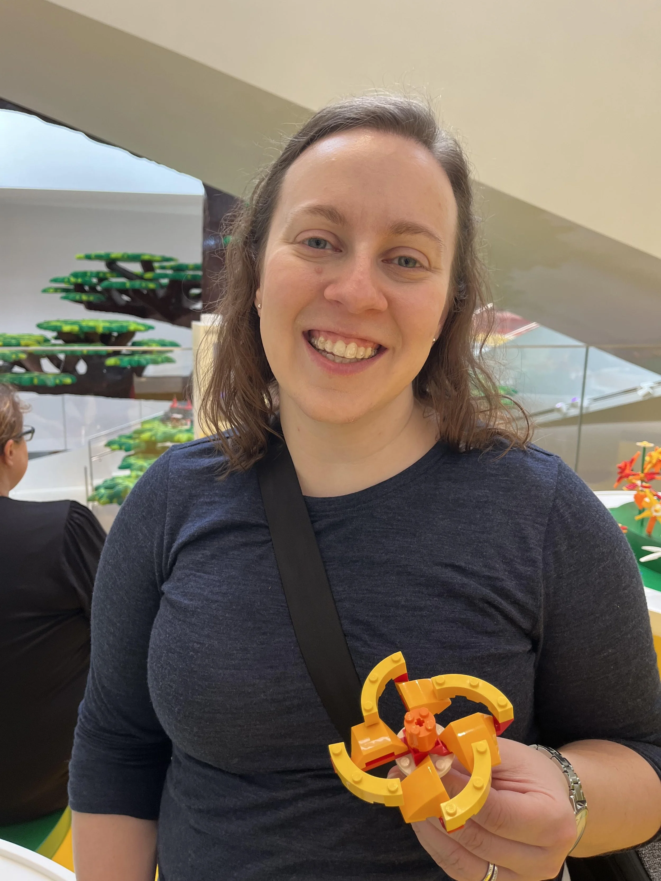 Woman smiling and holding a LEGO creation in an indoor setting with LEGO trees in the background.