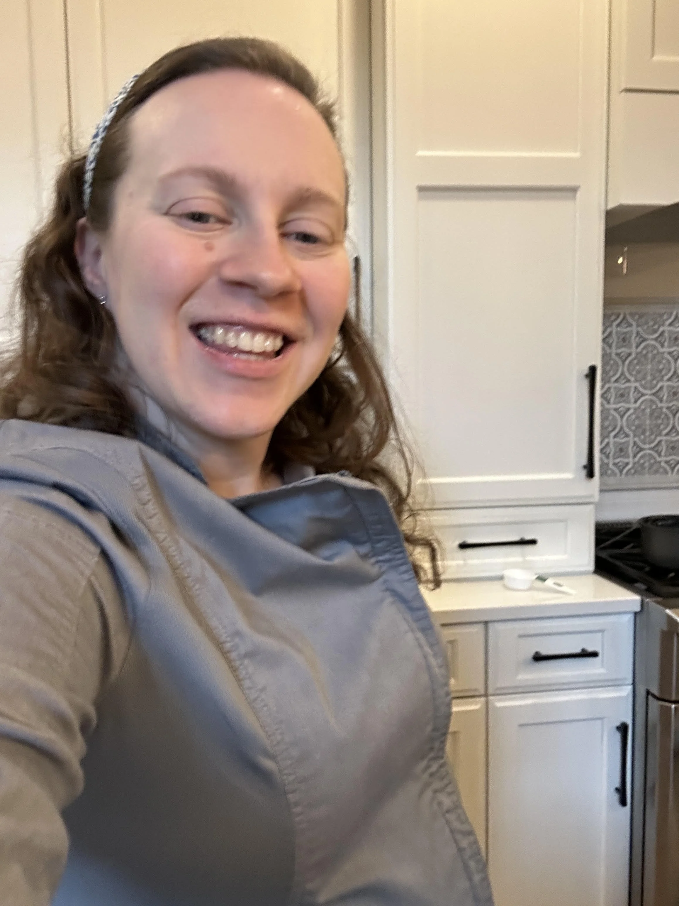 A woman smiling in a kitchen with white cabinets and black handles, wearing a gray jacket and a headband.