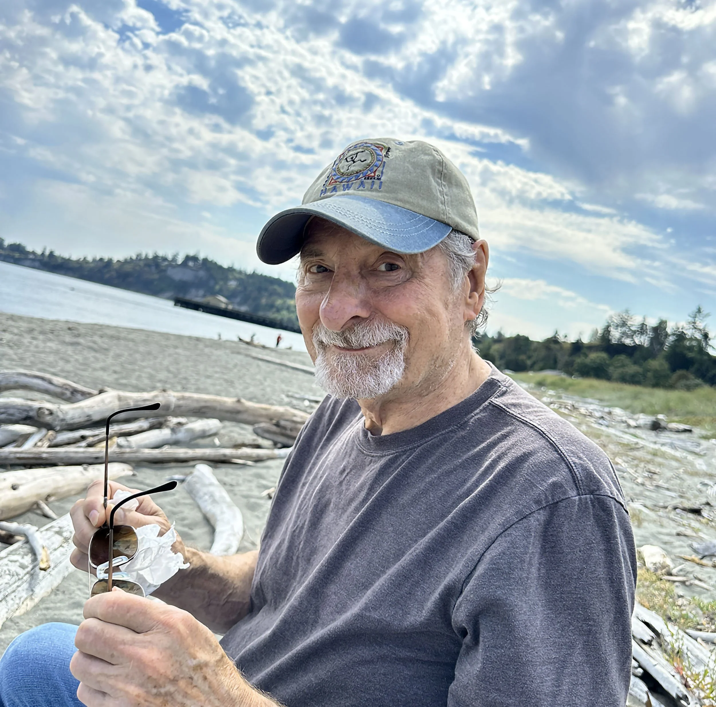 A smiling older man with a gray beard and mustache, wearing a beige and blue cap, sitting on a beach with pieces of driftwood, holding sunglasses in his hand. In the background, there is a sandy beach, water, and a partly cloudy sky.