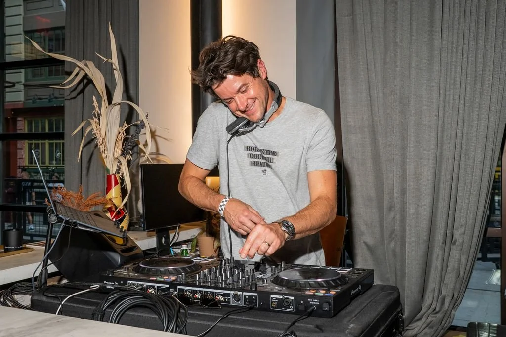 A man wearing a grey t-shirt and headphones around his neck is DJing with a Pioneer DJ controller, smiling and enjoying the music in a room with large windows, curtains, and decorative plants.