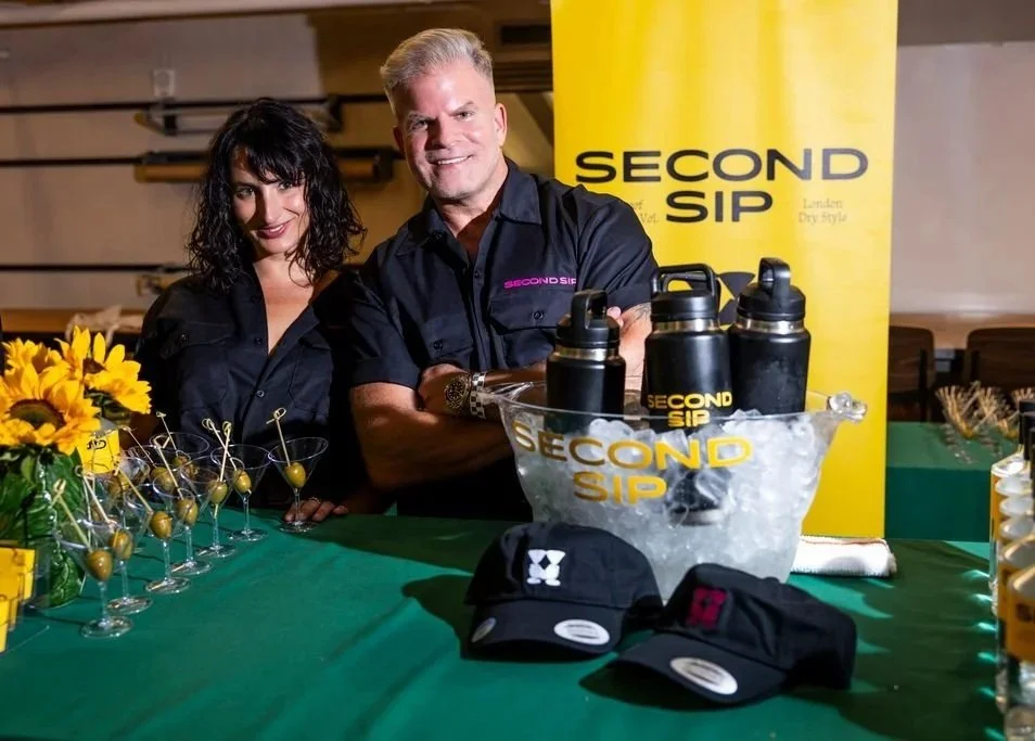Two people, a woman with dark wavy hair and a man with gray hair, standing behind a table with water bottles and glasses, at a promotional event for Second Sip, a beverage brand. There are yellow flowers on the left and a yellow banner with the brand