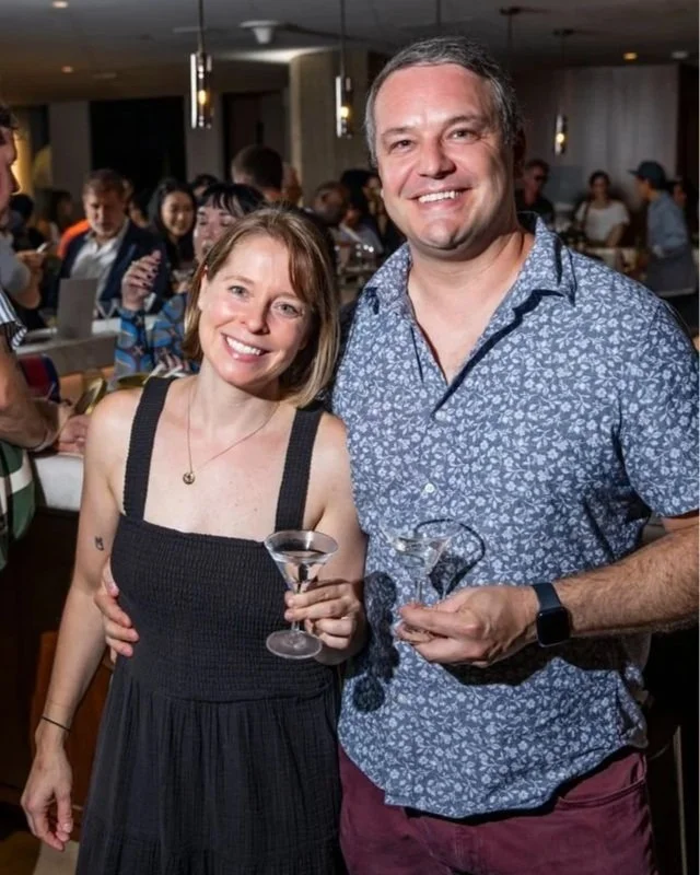 A smiling woman and man at a party, holding drinks, surrounded by a crowd in a lively indoor setting.