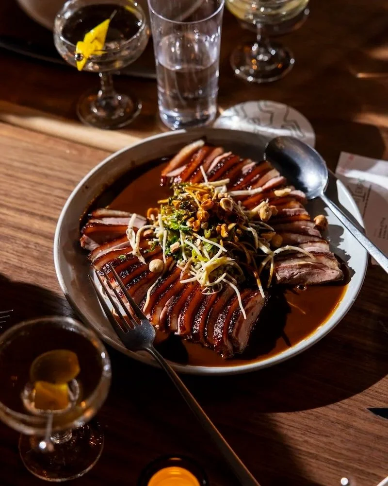 Dinner at Martini Expo New York: A plate of sliced duck breast with crispy skin, garnished with bean sprouts and chopped nuts, served in a dark sauce, with drinks and glass of water on a wooden table.