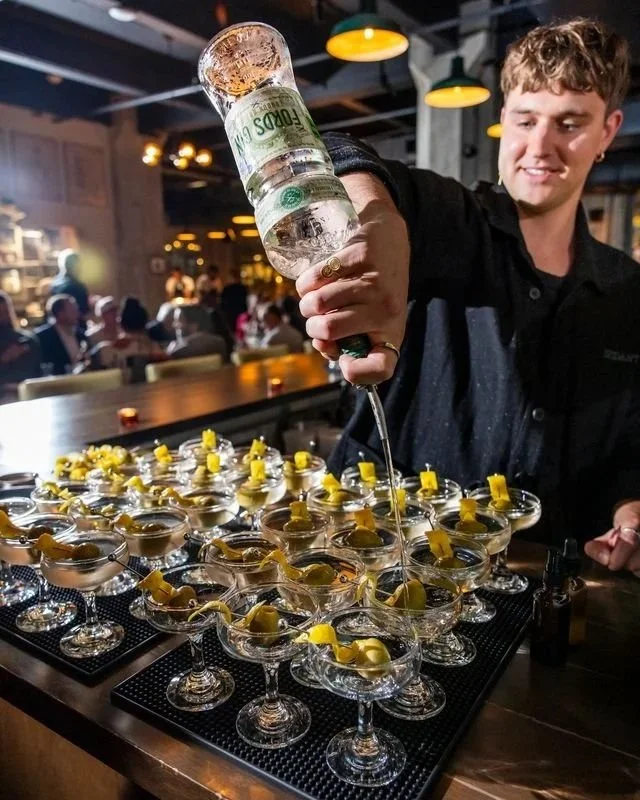 A man pours a large bottle of vodka into multiple cocktail glasses with lemon slices in a bar or restaurant setting.