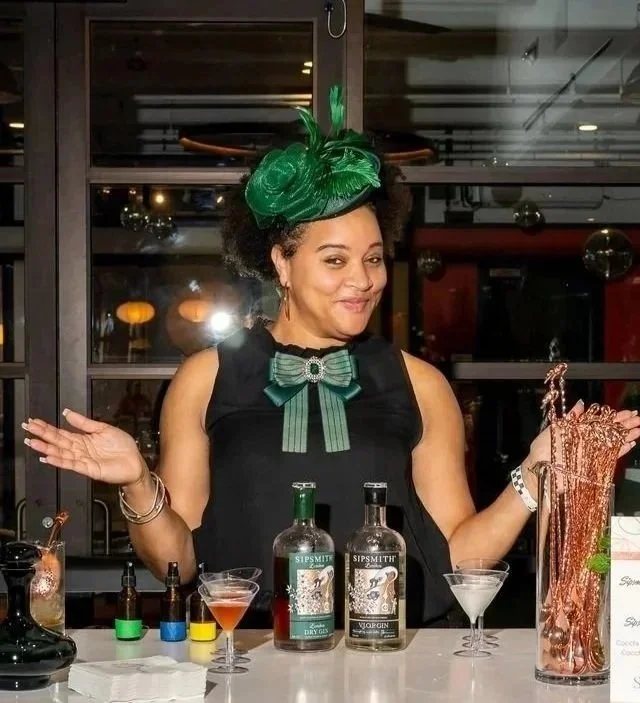 A woman in a black dress and green hat with a feather, standing behind a bar with alcohol bottles and cocktail glasses, smiling with arms open.