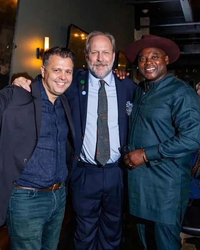 Three men smiling with arms around each other in a dimly lit indoor setting, possibly at a social event or gathering.