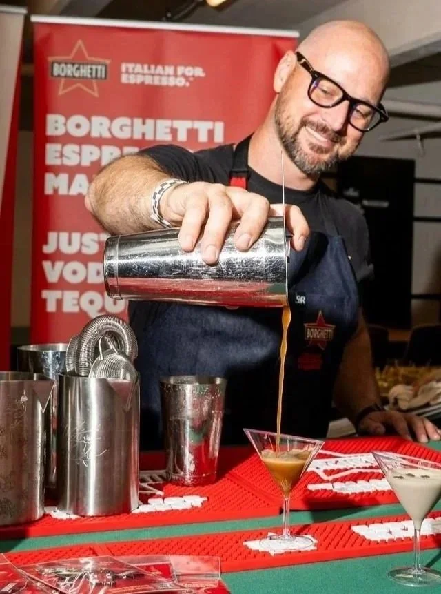 A bartender pouring a drink into a martini glass at a bar. The background features a red banner with white text promoting Italian espresso and other beverages. The bar counter has bar tools and napkins.