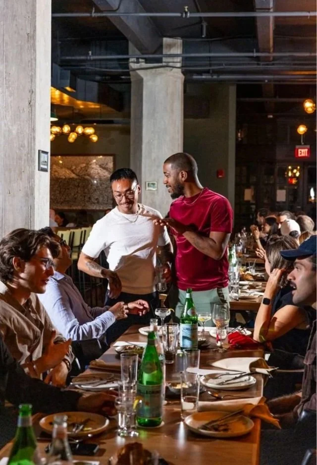 People enjoying a meal at a restaurant, seated around a table with plates, glasses, and bottles. Two men are standing and conversing, one wearing a white shirt and the other a red shirt.