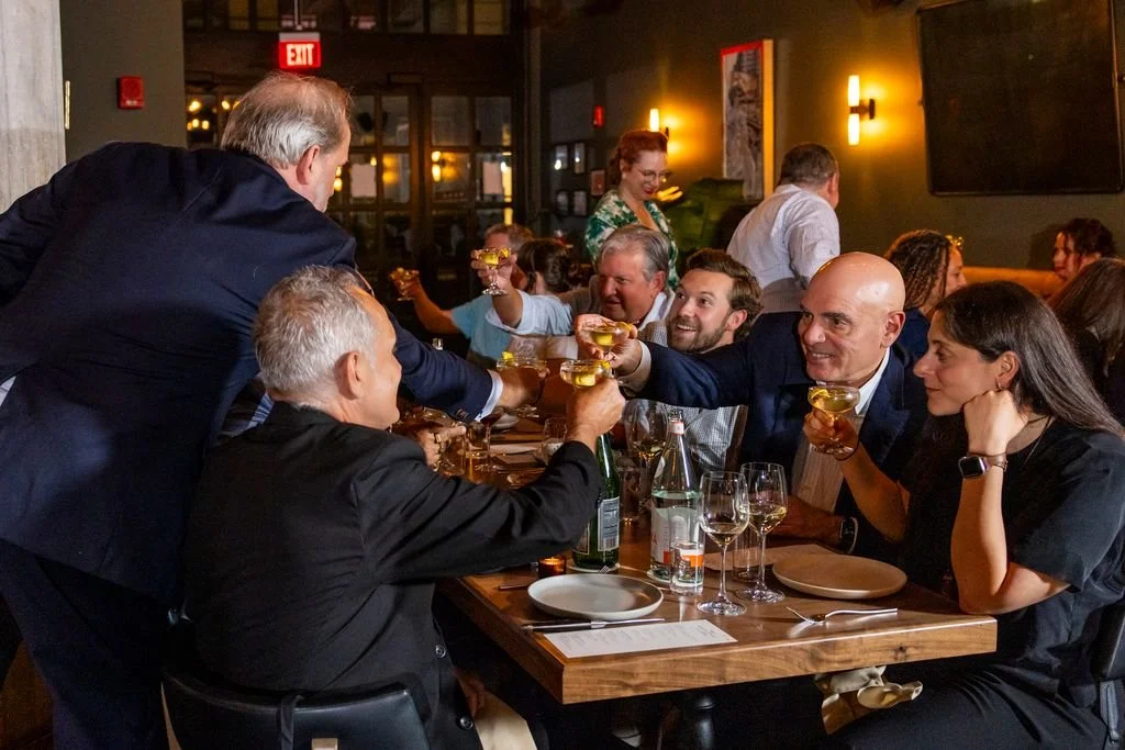 Group of people celebrating at a restaurant, raising glasses for a toast, with a server serving drinks to the table.