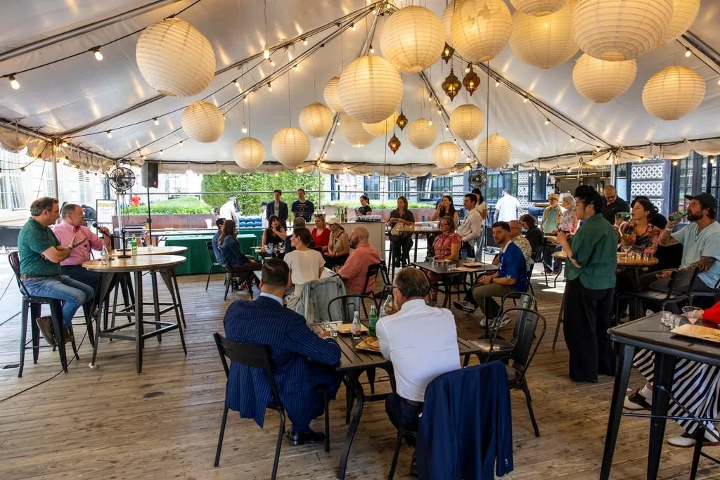 A group of people gathered under a white canopy at a restaurant or event space, with hanging paper lanterns and string lights, some seated at tables and others standing, during daytime.