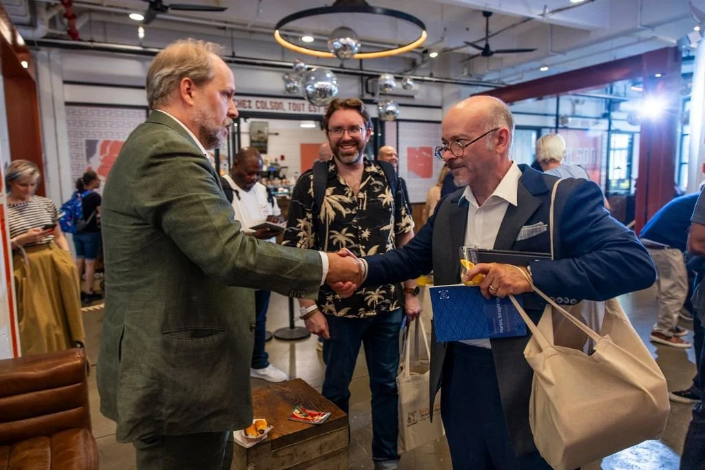 Two men shaking hands at an indoor event, with one holding a glass of drink and a bag, in a busy room with other people and a modern decor.