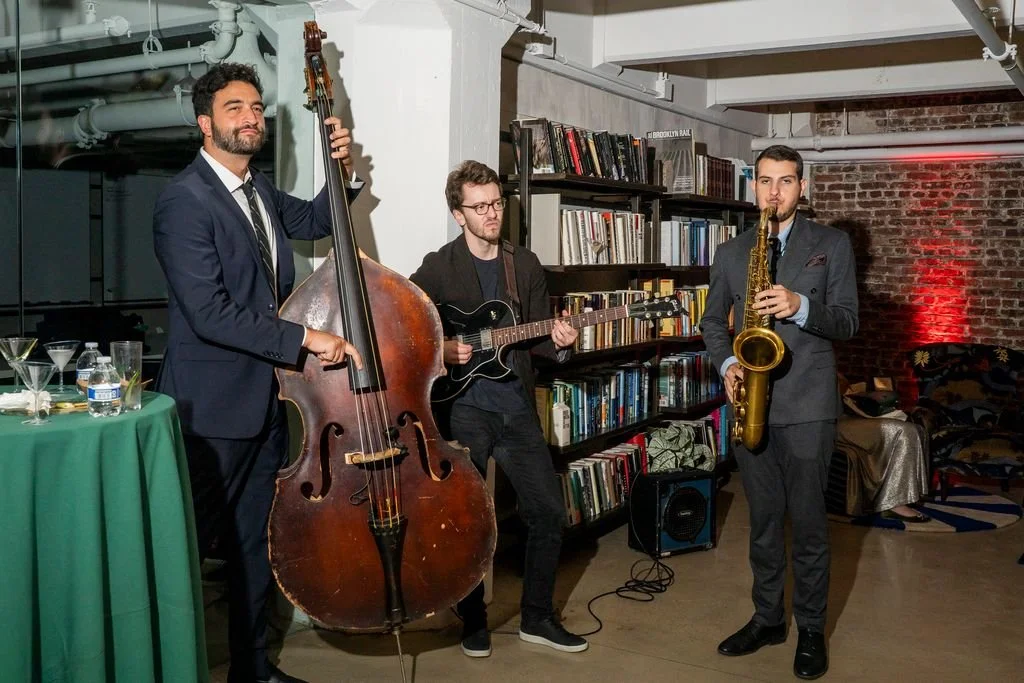 Three men in suits performing jazz music with a double bass, electric guitar, and saxophone in a cozy, modern space with brick walls and bookshelves.