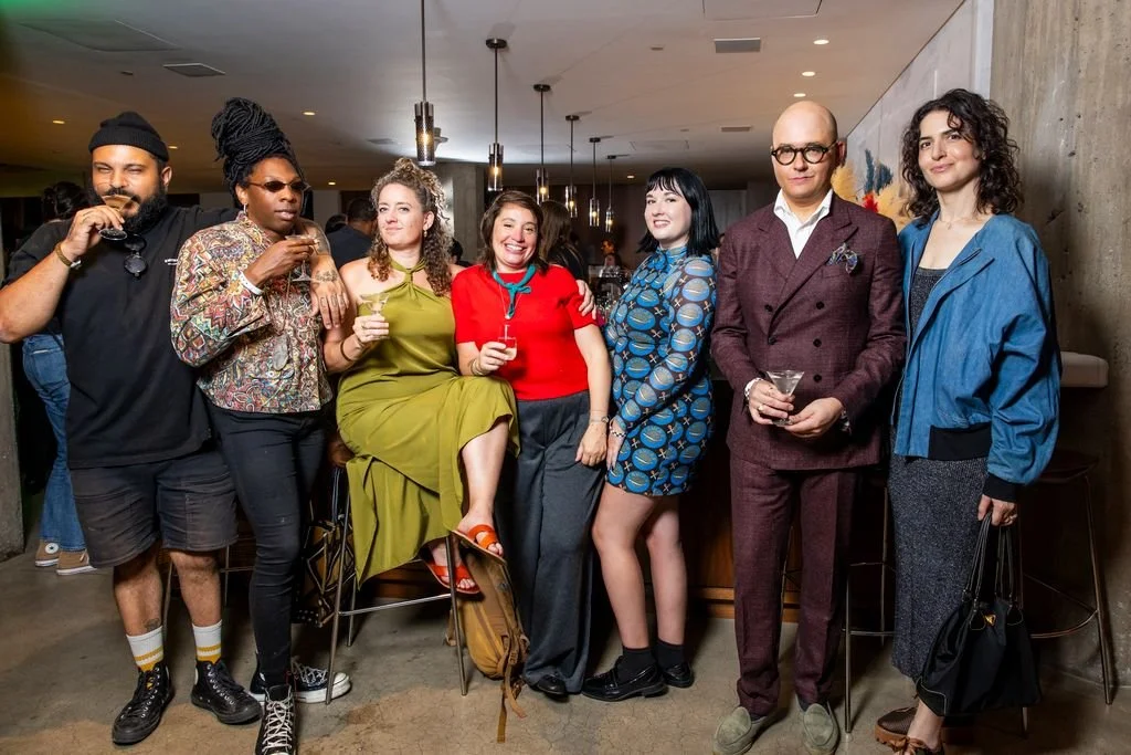 Group of seven diverse people at a social gathering, some holding drinks, smiling and posing for the photo in an indoor setting.
