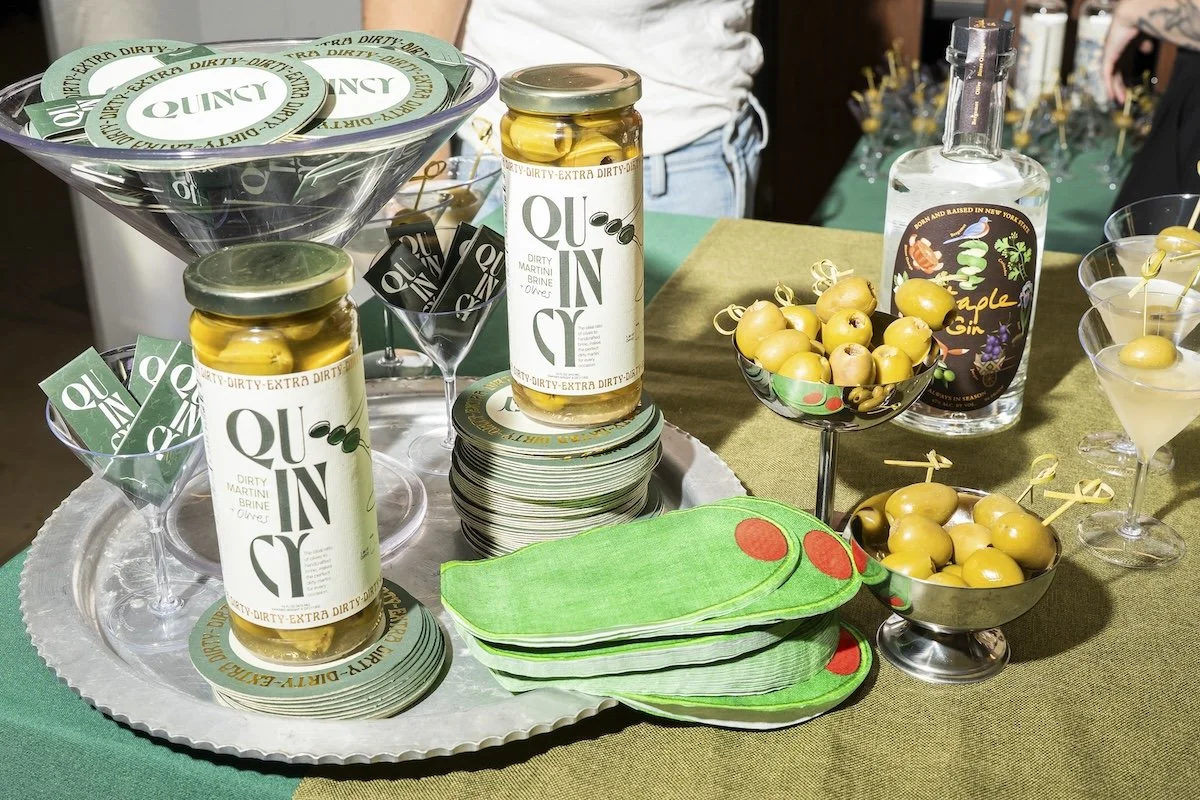 A table set with Quinte branded jars of pickled olives, small bowls of yellow olives on cocktail picks, martini glasses with stuffed green olives, a bottle of apple gin, green and red napkins, and a large glass bowl with more stuffed olives, all arra