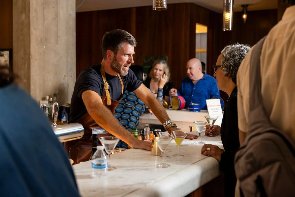 Bartender leaning on bar counter, serving drinks to customers in a lively bar setting with wooden walls and hanging lights.