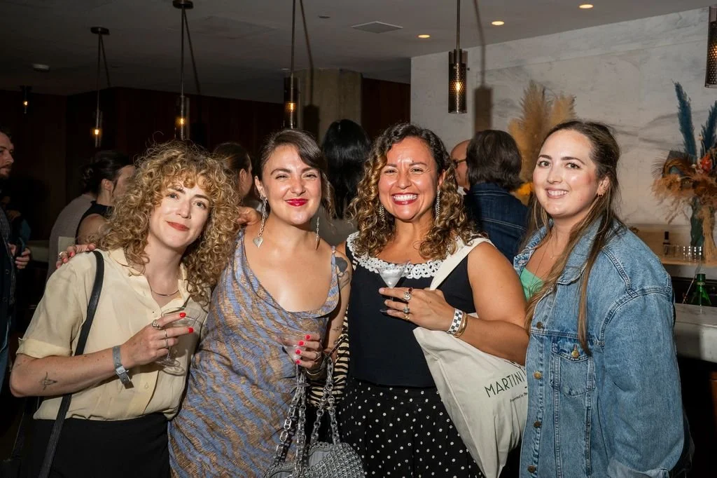 Four women smiling at a social event, with drinks in hand, in a well-lit indoor setting.