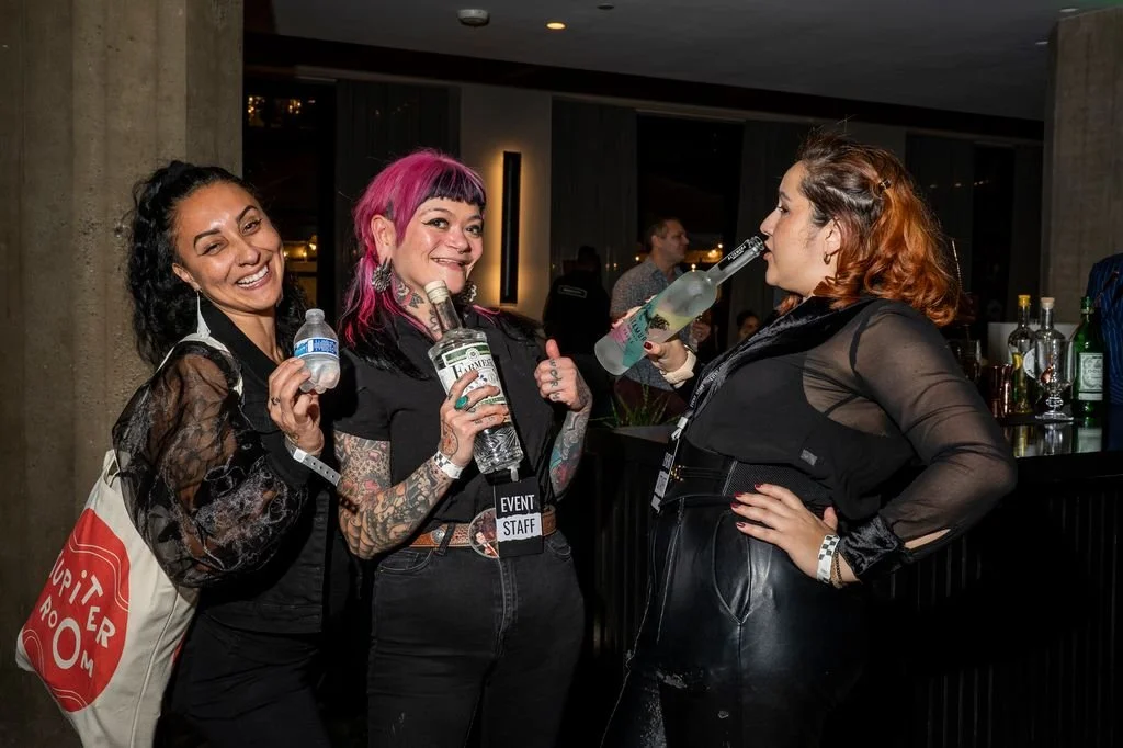 Three women at a social event, two holding bottles and one drinking from a bottle, standing near a bar with drinks in the background.