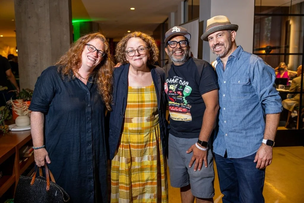 Four people standing close together, smiling, indoors at a social event. There are plants and people in the background.