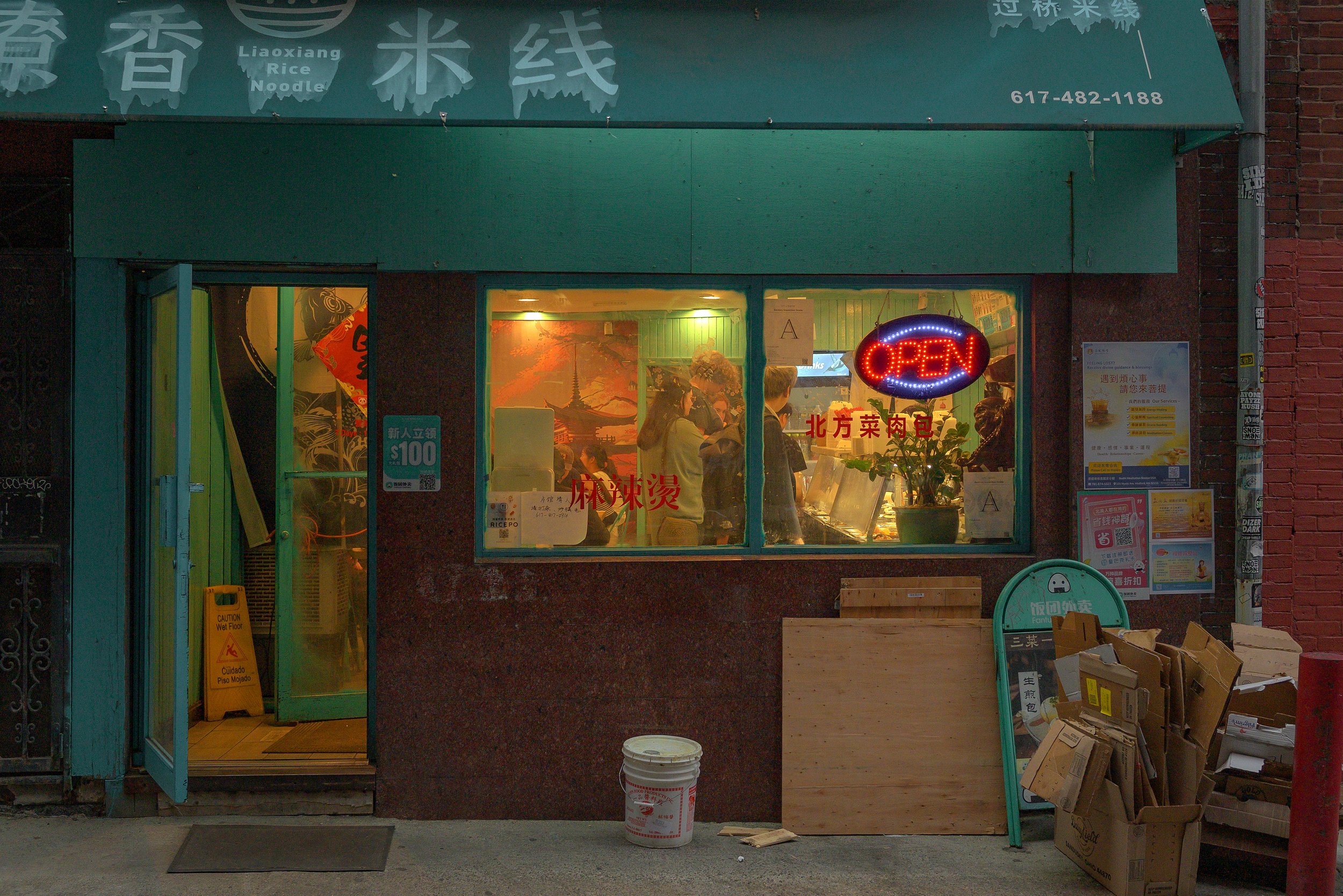 A small restaurant with a green roof displaying a neon 'OPEN' sign in the window. Inside, there are people dining. The storefront has Chinese characters and English words reading 'Liaoxiang Rice Noodle'. There are various posters and signs outside th