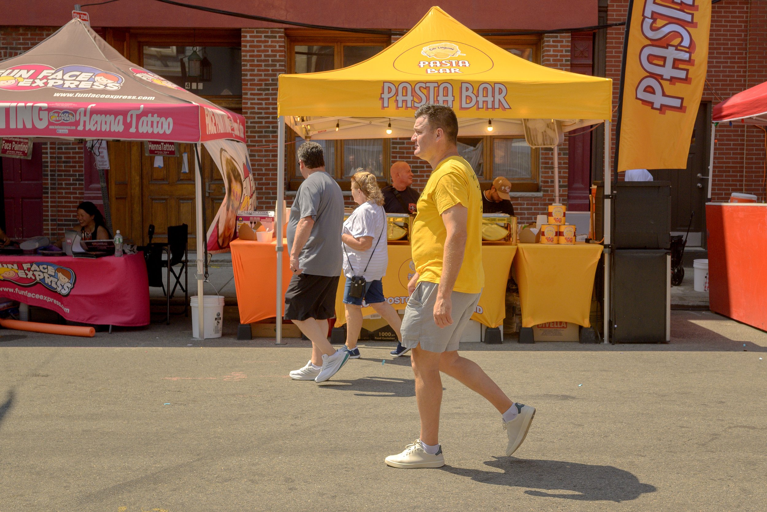 People walking past food stalls at an outdoor event with brick buildings in the background. One stall is yellow with a sign for a pasta bar, and another is pink advertising tattoos and face painting.