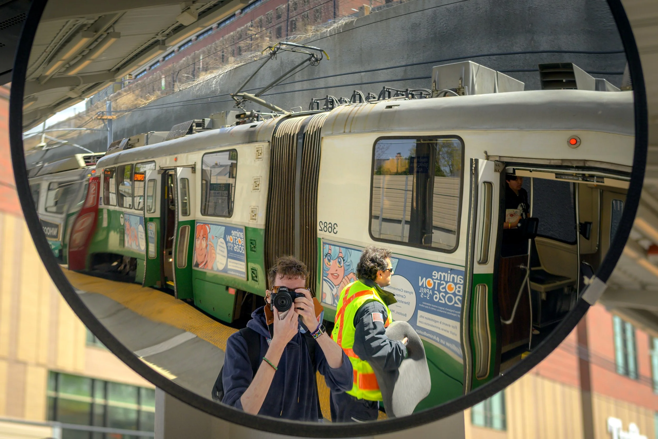 Reflection of a person taking a photo of a subway train and a person in a yellow safety vest in a circular mirror.