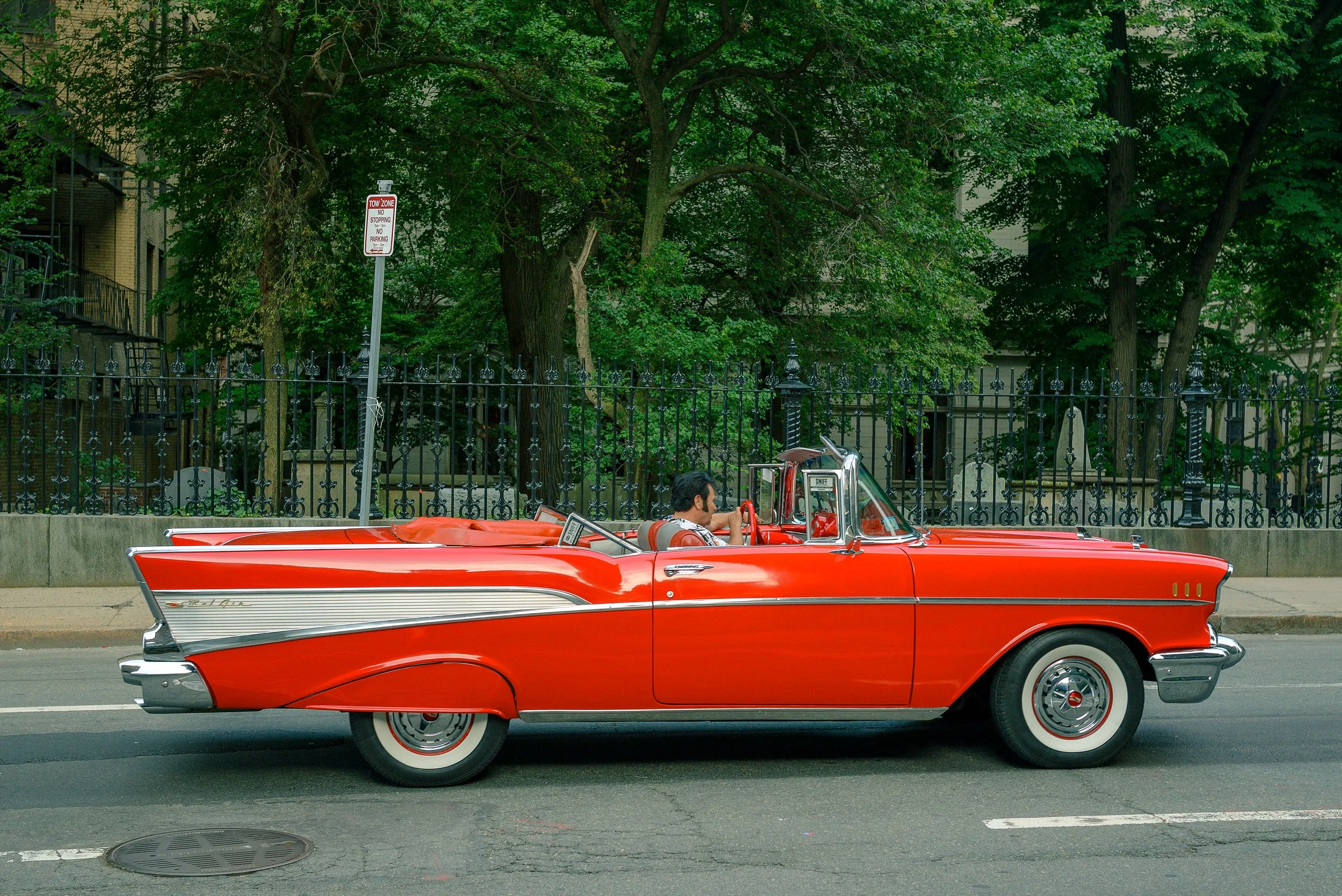 A vintage red convertible car with a man driving on a city street, with green trees and a black wrought iron fence in the background.