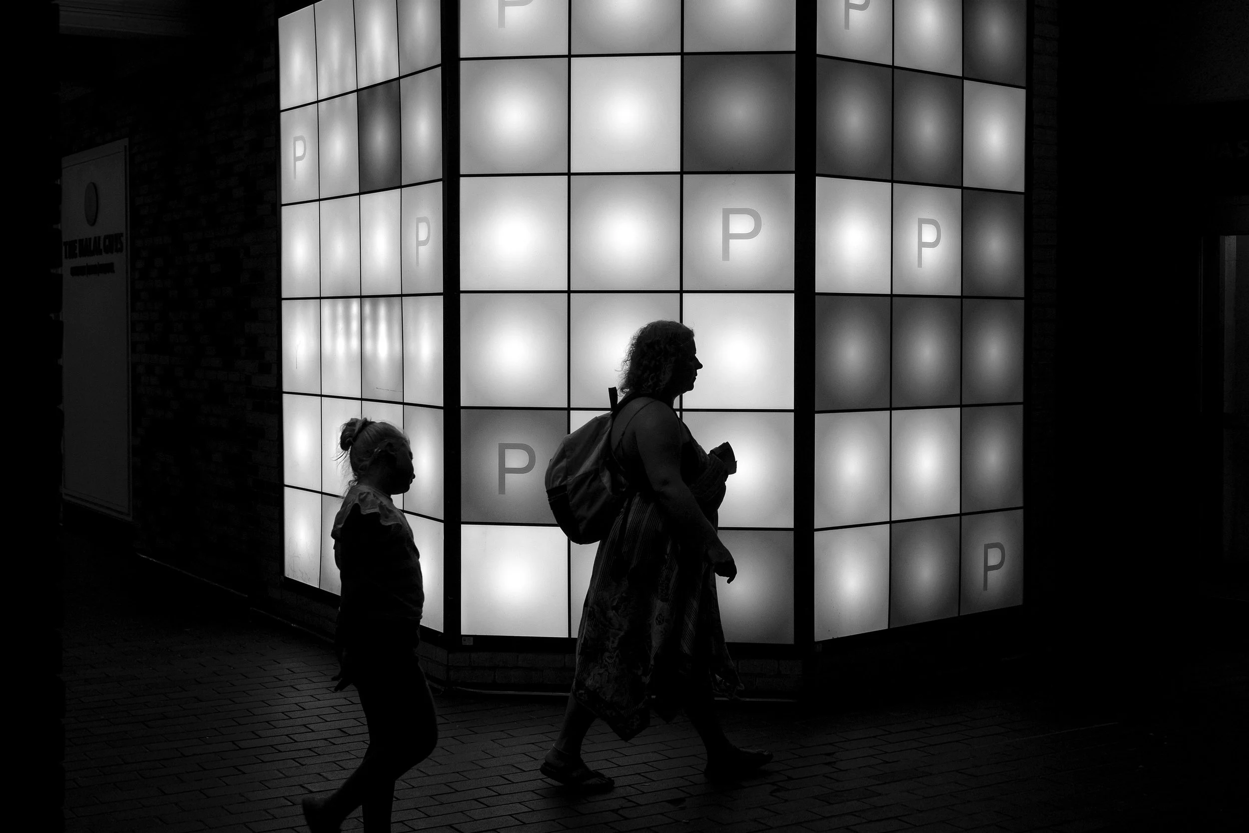 People walking past a building with illuminated panels displaying the letter 'P' in black and white.