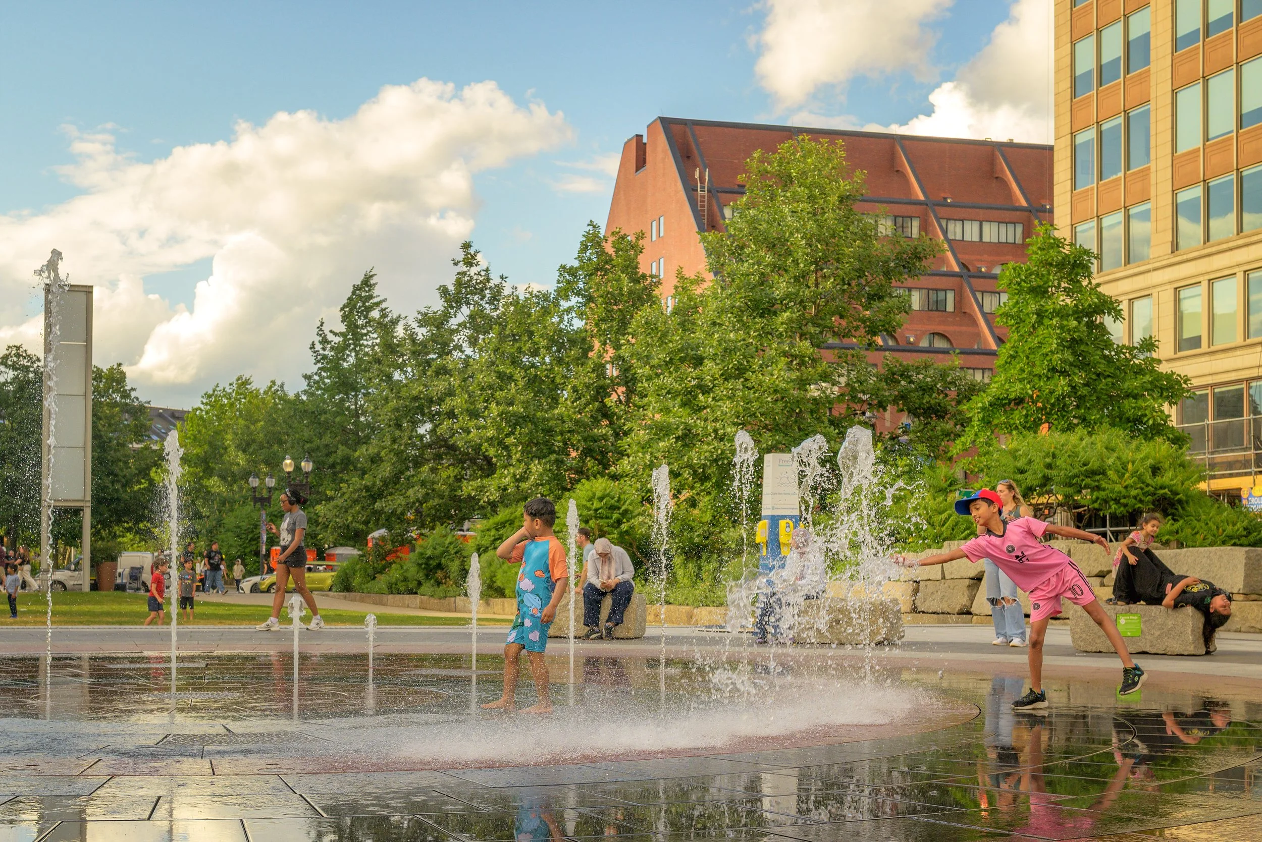 People playing and splashing in water fountains in an urban park with tall buildings and trees, under a partly cloudy sky.