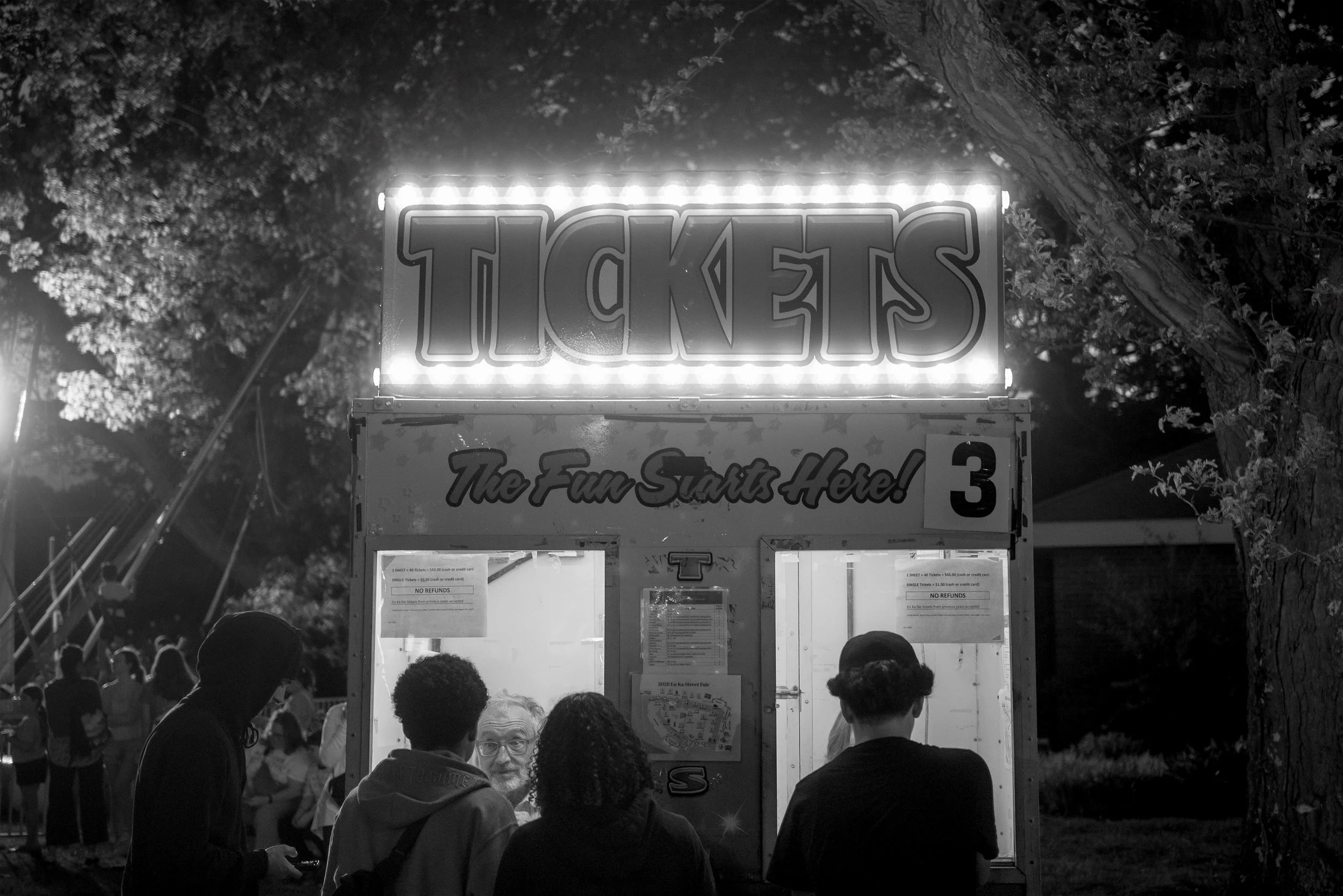 A black and white photo of a ticket booth at a fair with a crowd of people in front, illuminated sign reading 'TICKETS' at night.