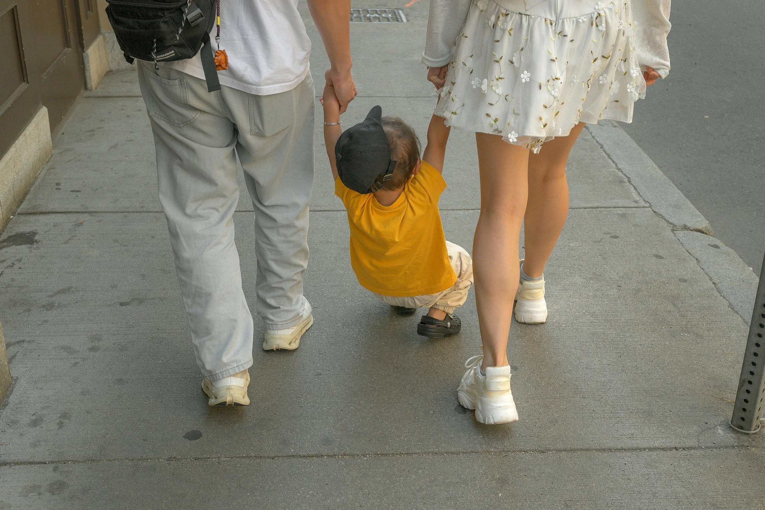 A young child with a yellow shirt and beige pants squatting on the sidewalk, holding hands with a woman wearing a floral dress and white sneakers, and an adult man wearing light-colored jeans and a white shirt, holding the child's other hand.