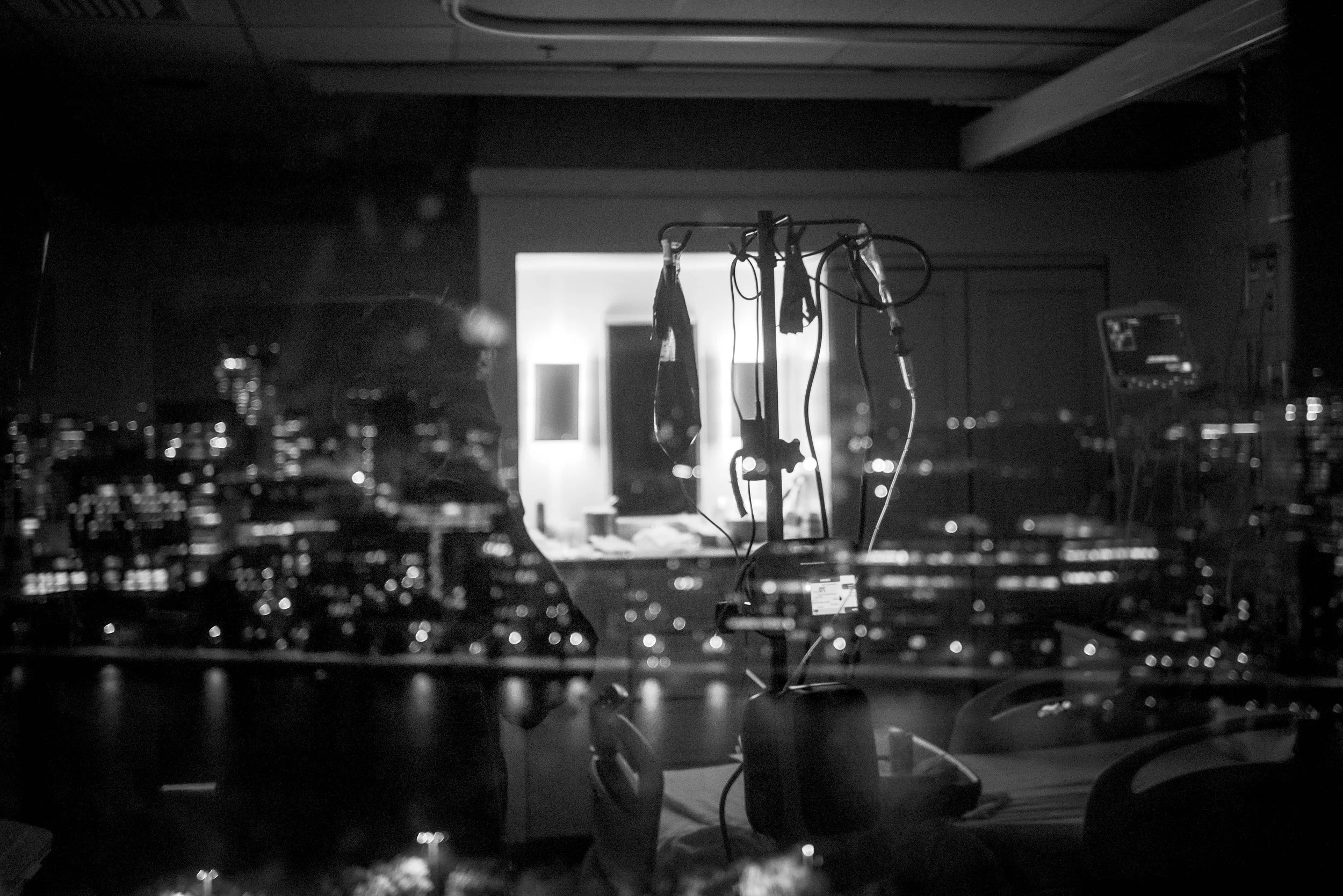 A black and white photo of a dimly lit room with various electronic equipment and wires. In the background, there is a window with bright light shining through, and objects hanging from a rack, possibly medical or electronic instruments.