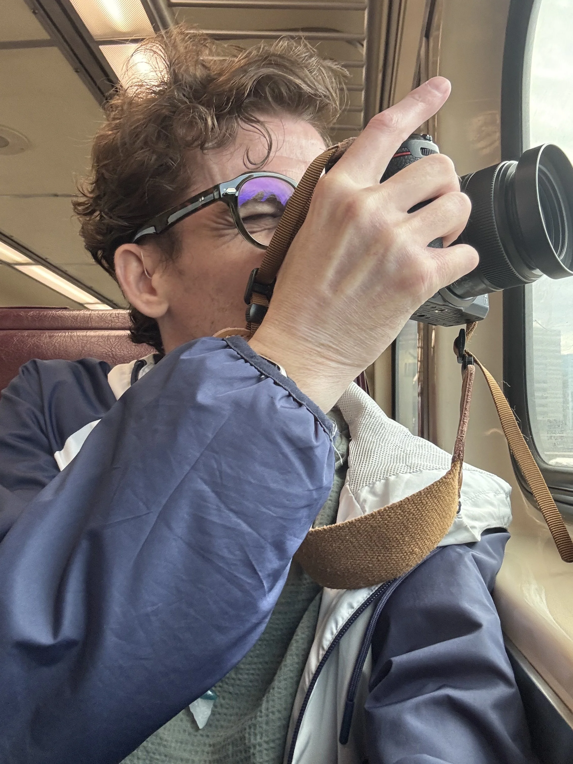 a curly haired man wearing a light windbreaker jacket and glasses holds a Nikon mirrorless camera up to his eye to take a photo out of the trains window