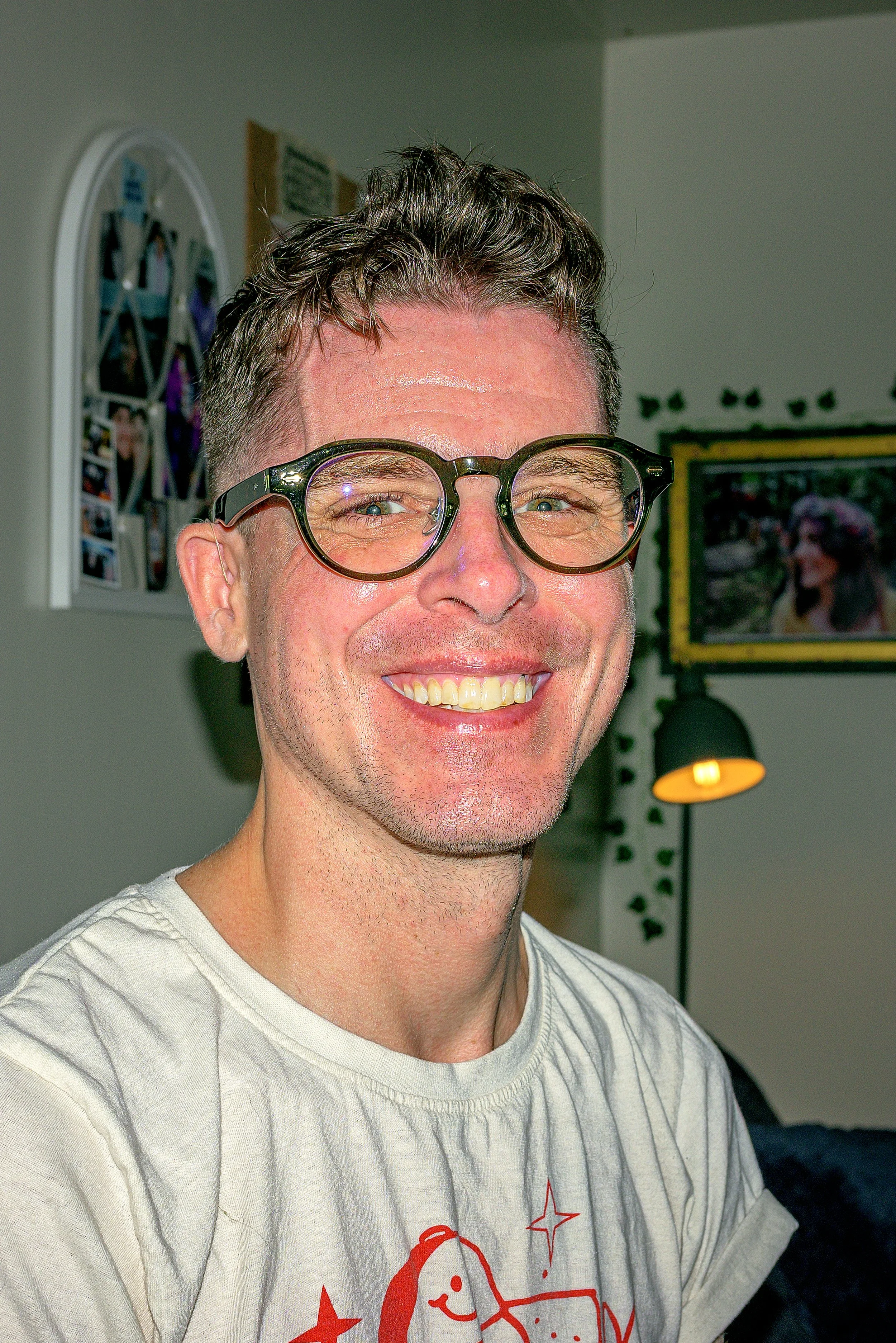 A smiling man with short dark hair and glasses wearing a white T-shirt with a red graphic design.