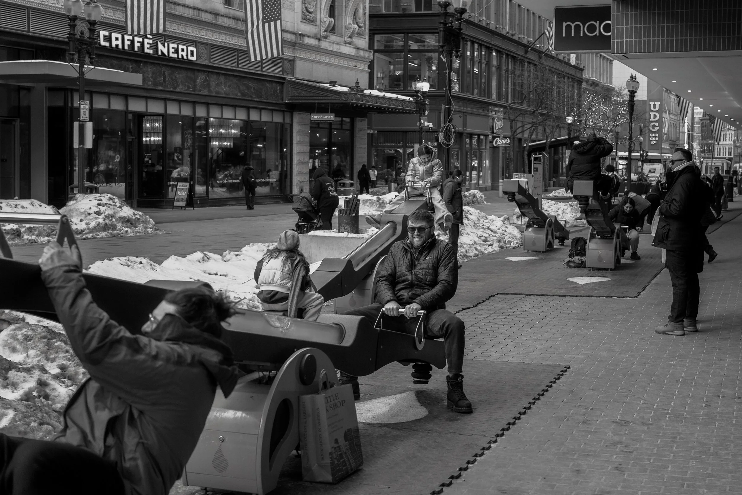 A lively city street scene in winter with people walking and enjoying an outdoor shopping area. Snow is piled along the sidewalk, and various storefronts are visible, including a cafe and retail stores. People are sitting on benches and playing aroun