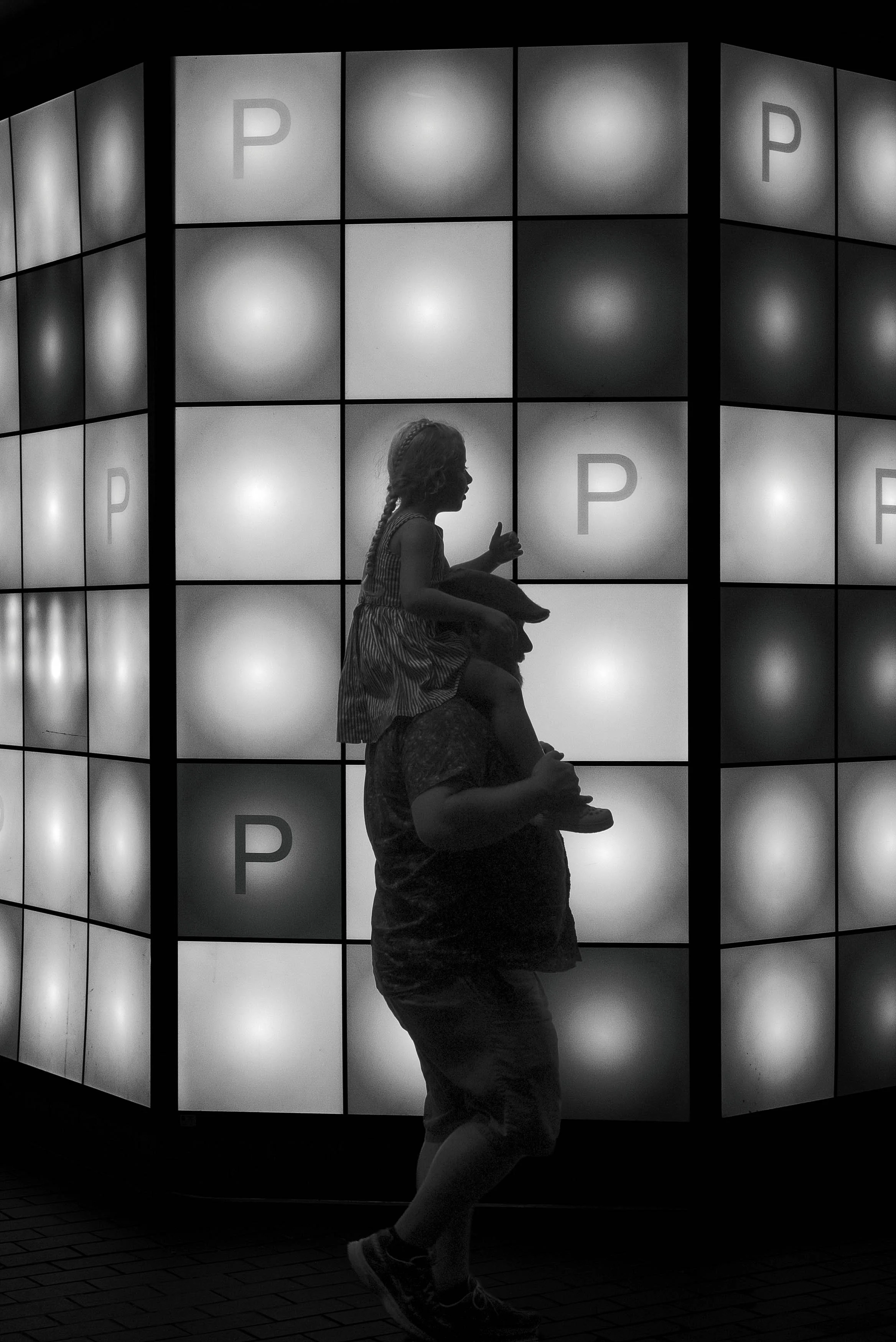 A silhouette of a young girl sitting on a man's shoulders in front of a large illuminated wall with multiple screens displaying the letter P. The girl appears to be giving a thumbs-up gesture.