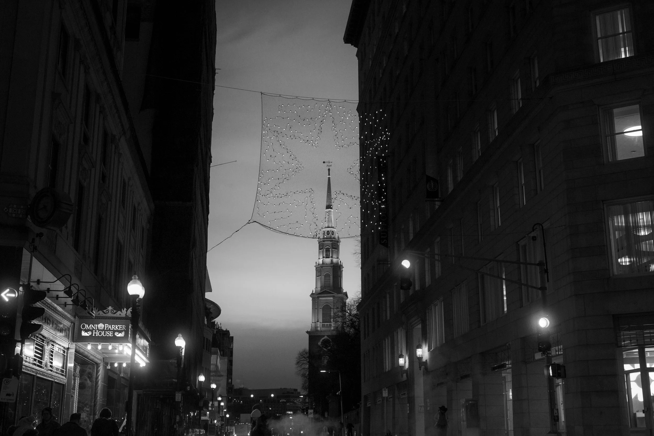 A narrow city street at dusk with hanging decorative lights, a church steeple in the background, and illuminated storefronts and street lamps on either side.