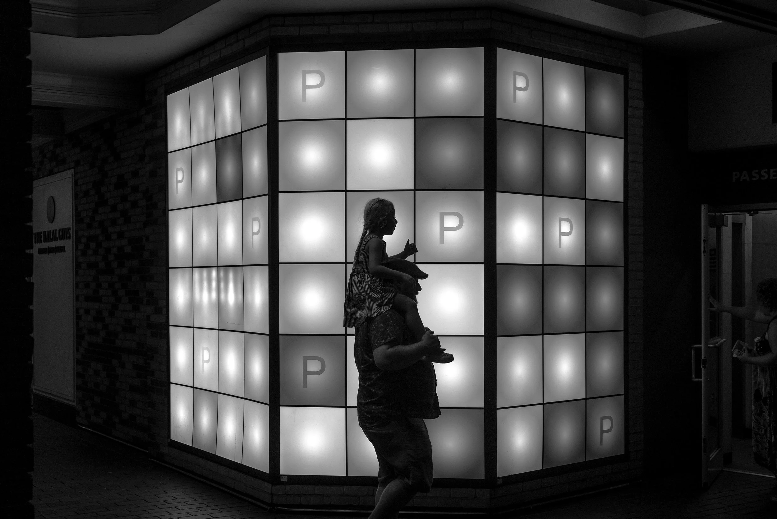 A black and white image of a girl sitting on a ledge and a man walking past, illuminated by a large lit-up window display with the letter 'P' on it.