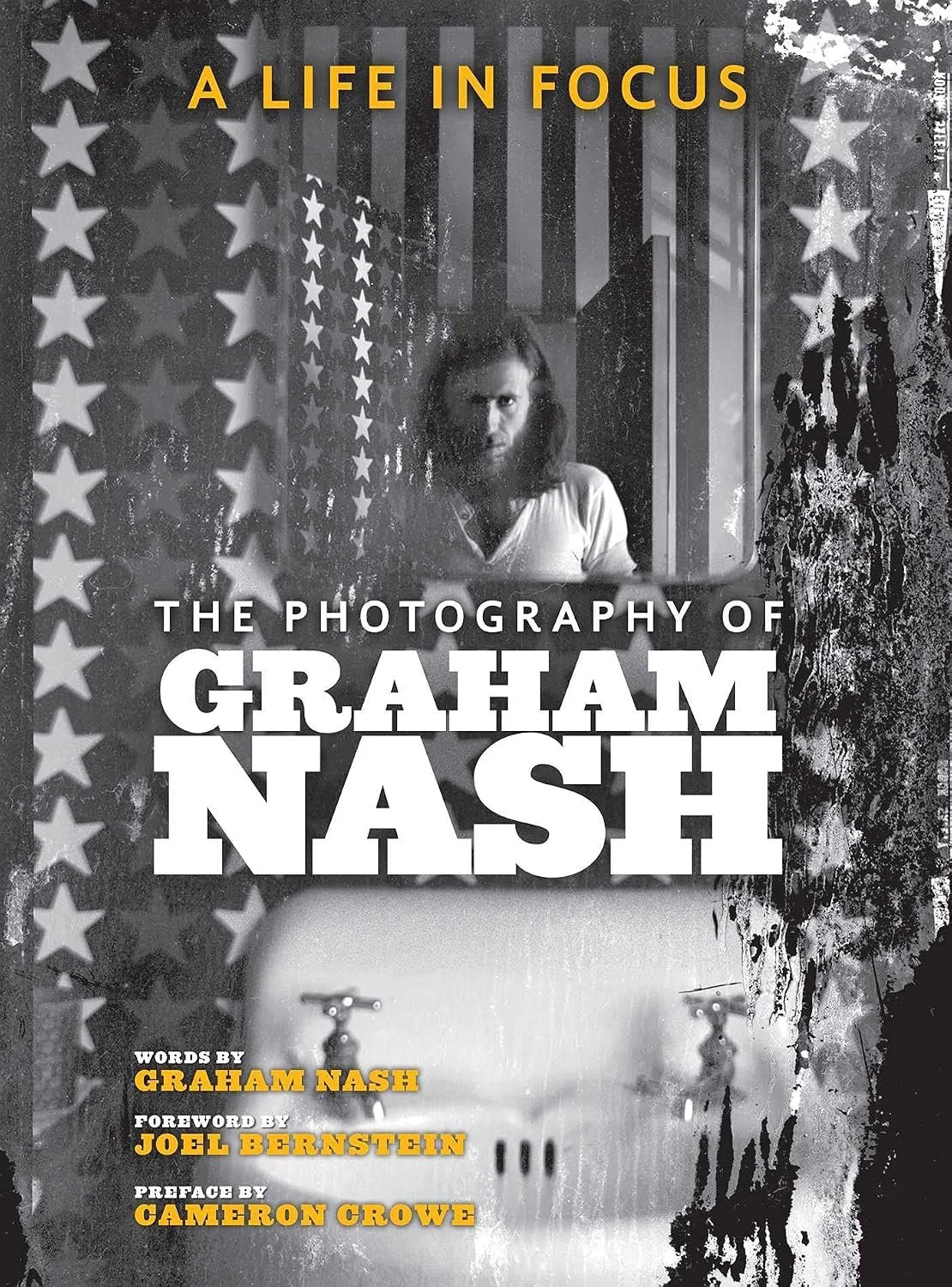 Book cover titled 'A Life in Focus' with a black-and-white photograph of a man with long hair and a beard looking into a mirror. The background features American flag motifs and a distressed, textured design. The cover includes the subtitle 'The Photography of Graham Nash' and credits words by Graham Nash, foreword by Joel Bernstein, and preface by Cameron Crowe.