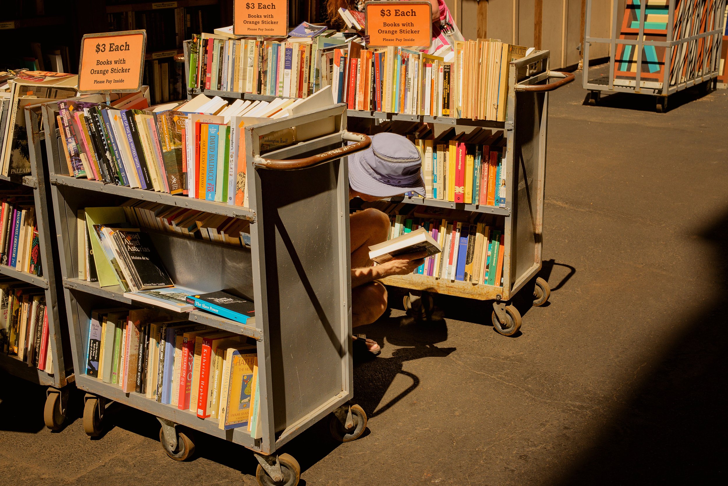 Outdoor book sale with multiple shelves filled with books and orange signs advertising books for $3 each with orange stickers, a person browsing books, and a person sitting on the ground reading a book, with a gray hat on the shelf.