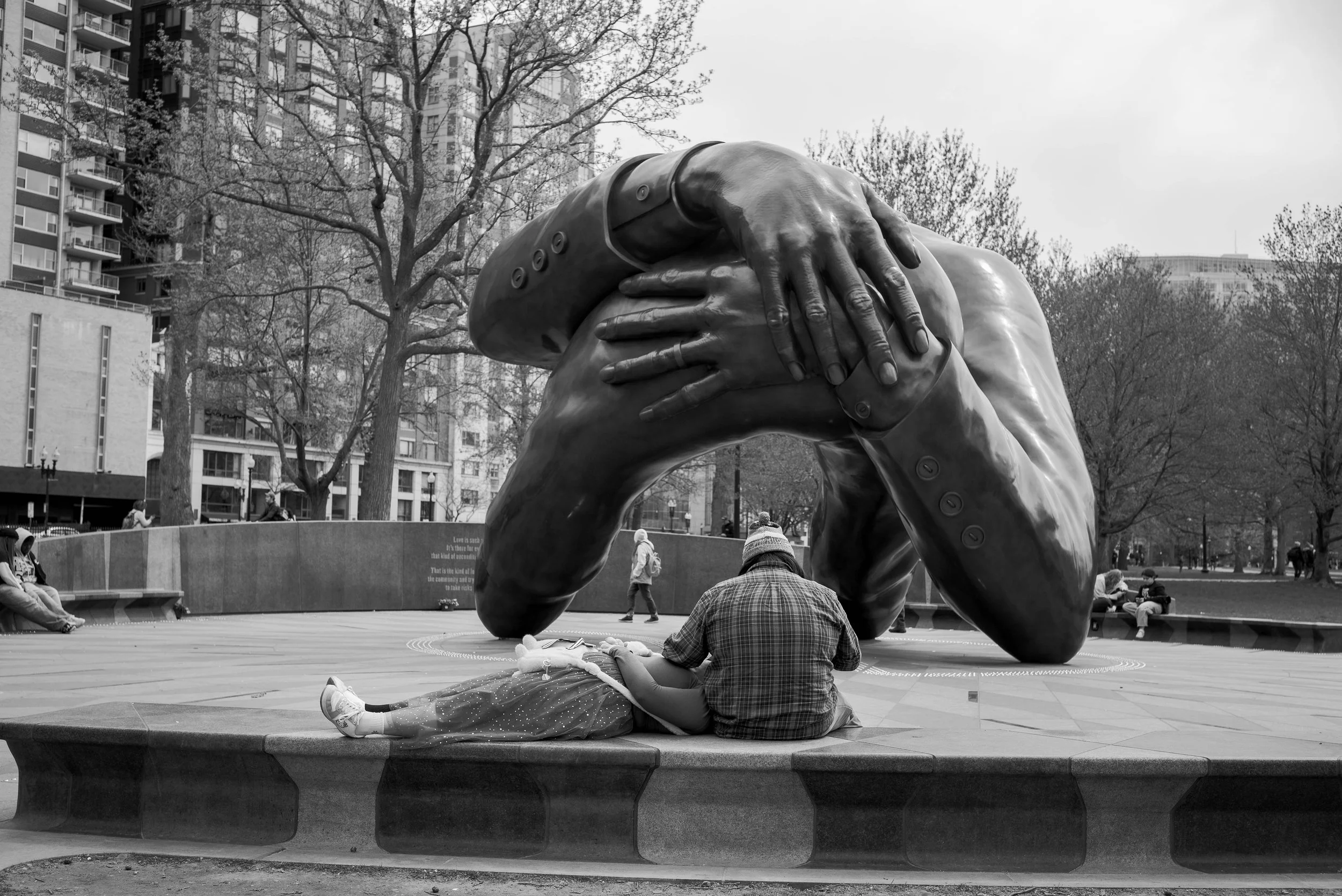 A large sculpture of a man with his head resting on his hand, situated in an urban park with trees and buildings in the background. Several people are sitting and walking around the sculpture, some reading or relaxing.