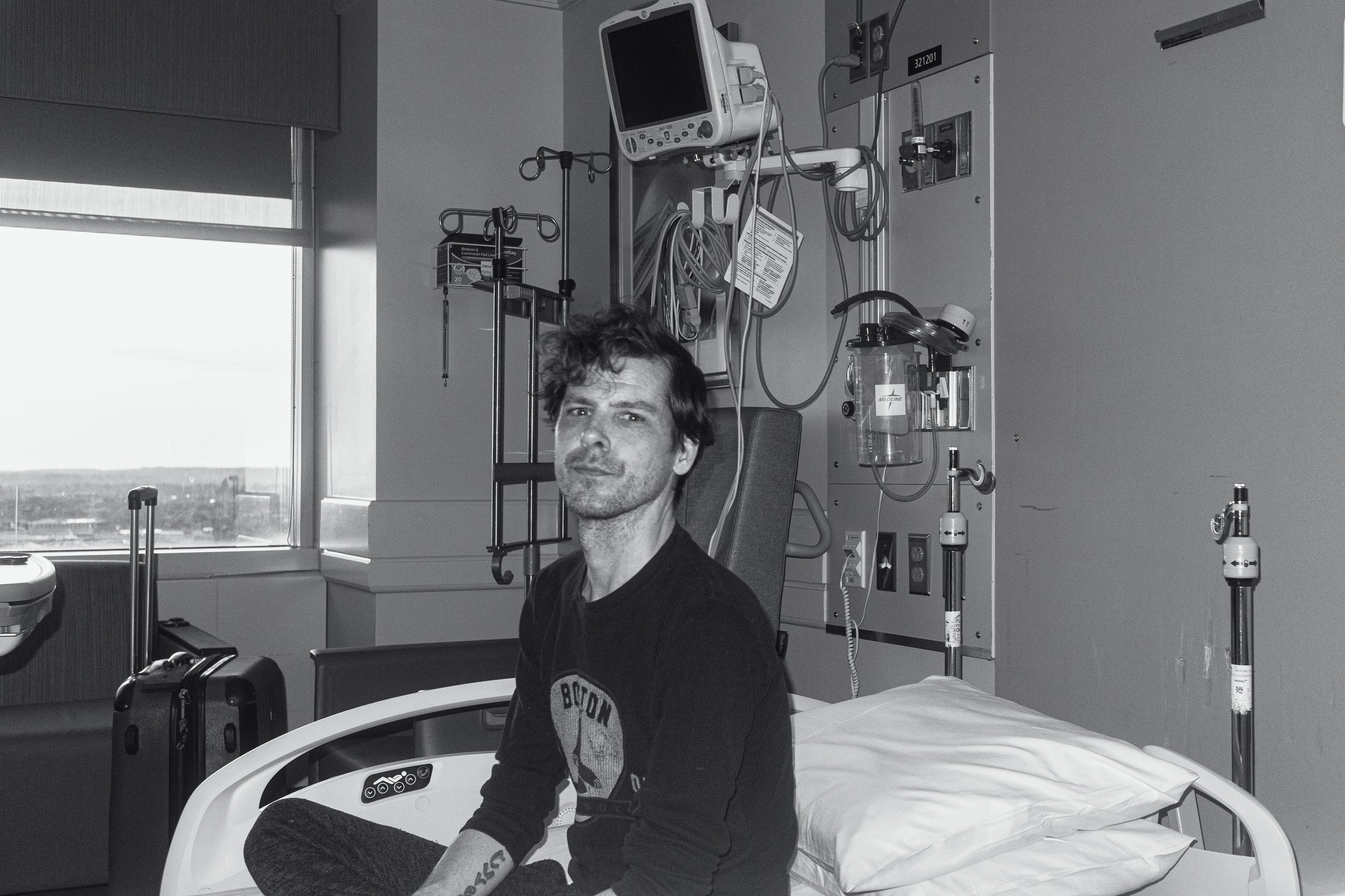 A man sitting on a hospital bed in a medical room with various equipment, including monitors, IV stands, and medical supplies, visible in the background, while looking at the camera.