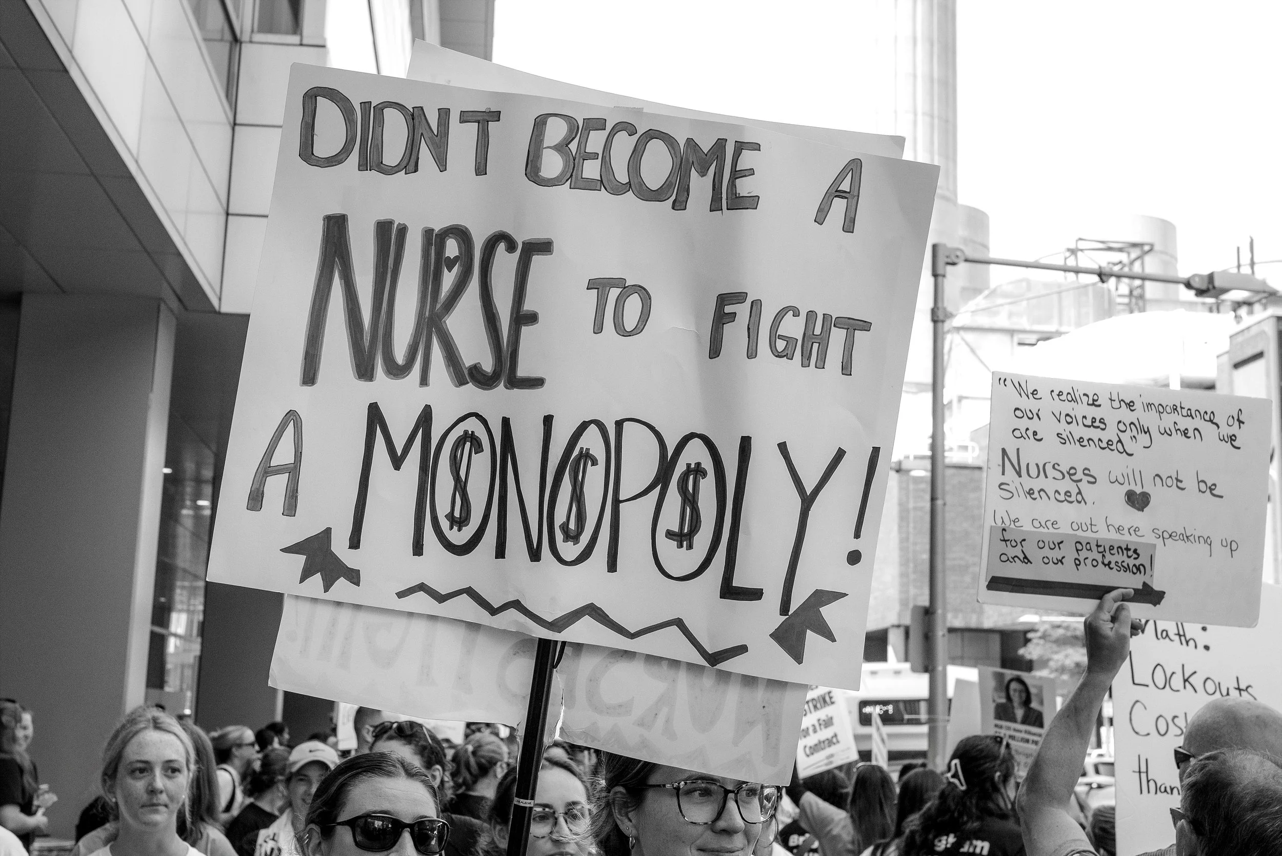 People at a protest holding signs, one sign says 'DONT BECOME A NURSE TO FIGHT A MONOPOLY!' a crowd in an urban setting with buildings and scaffolding visible in the background.