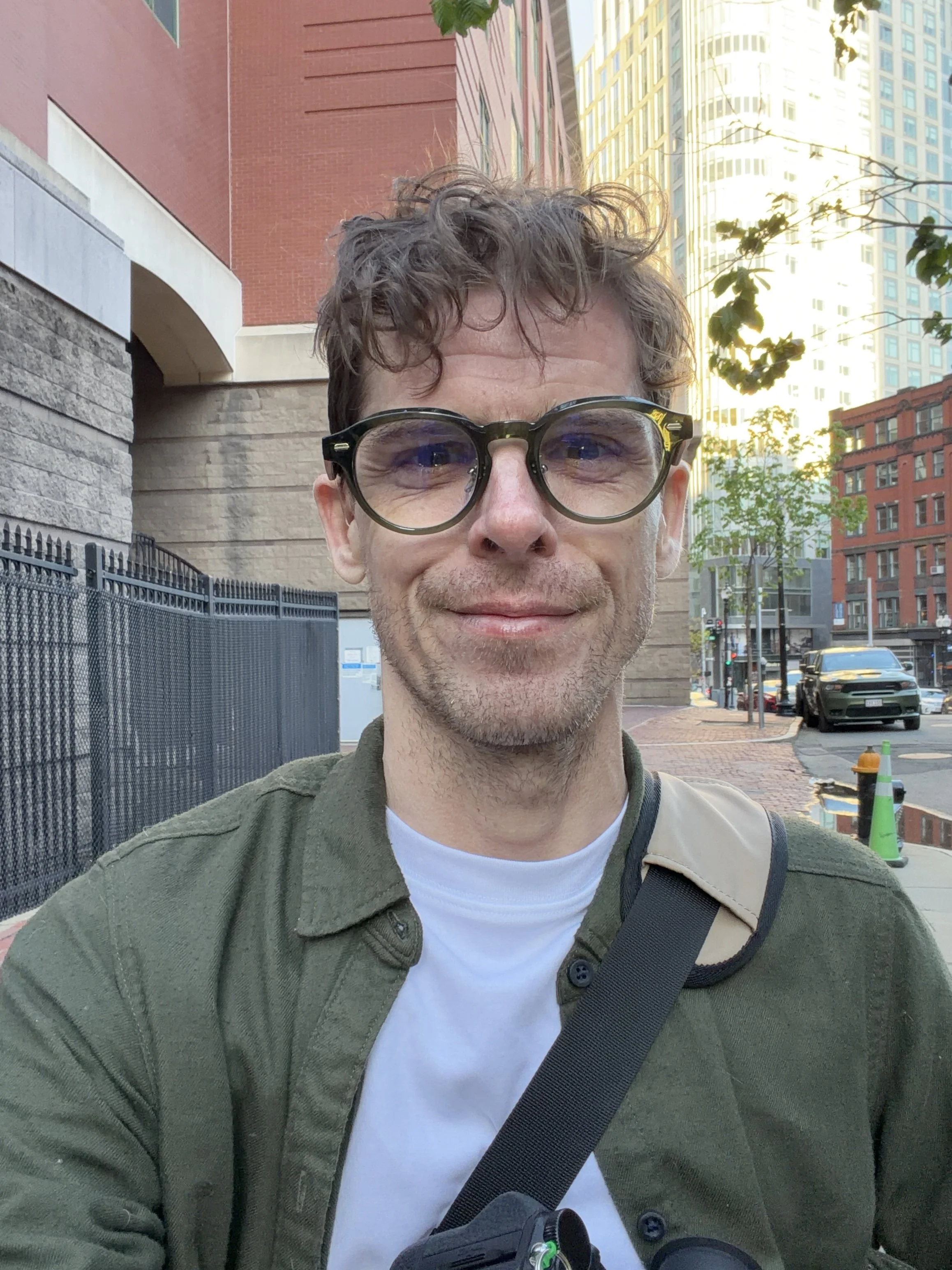 A man with curly hair and glasses smiling, walking in an urban area with tall buildings, a fence, trees, and parked cars.