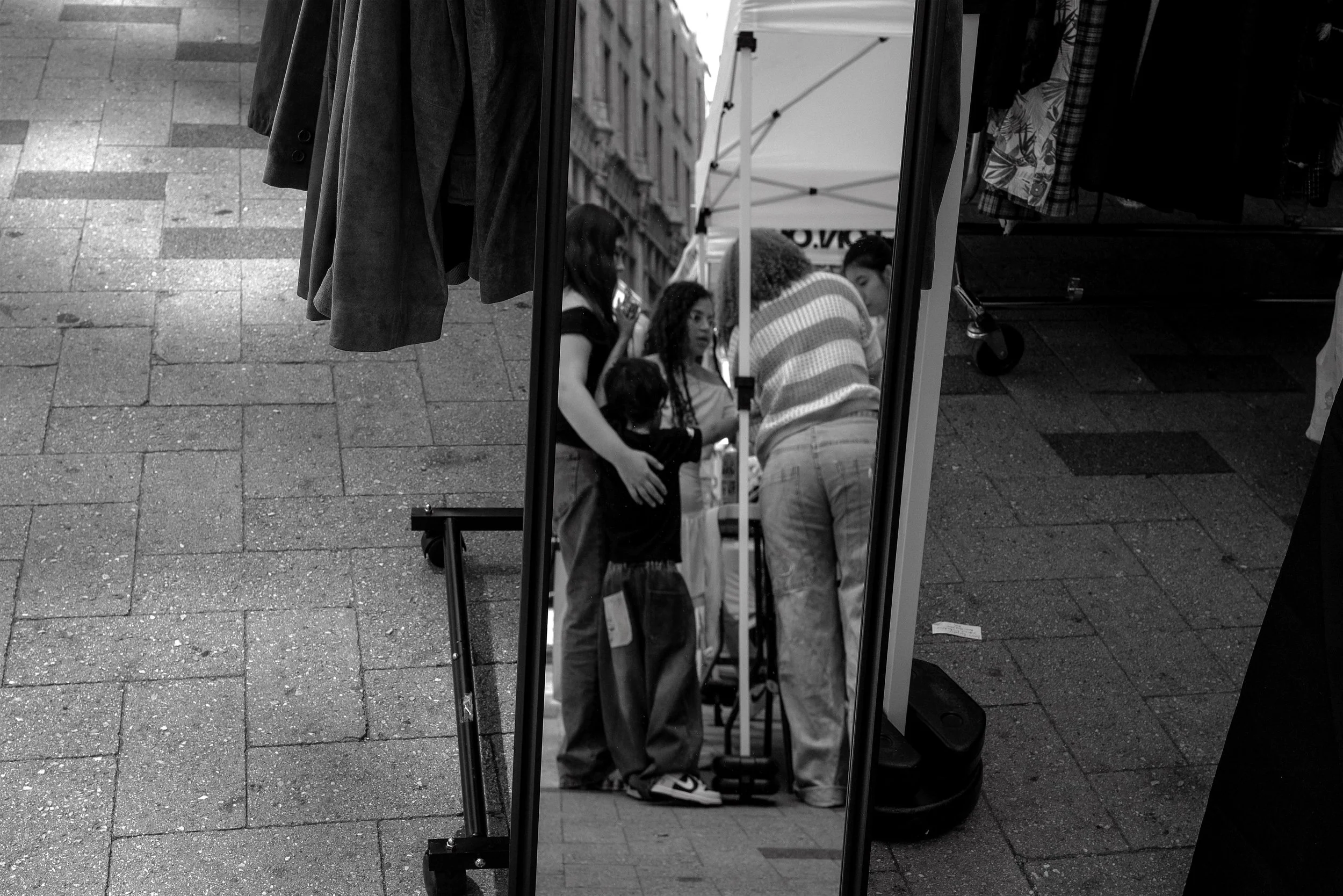 Street scene showing a row of outdoor clothing on display, with a mirror reflecting three young women browsing and talking, and a building in the background.