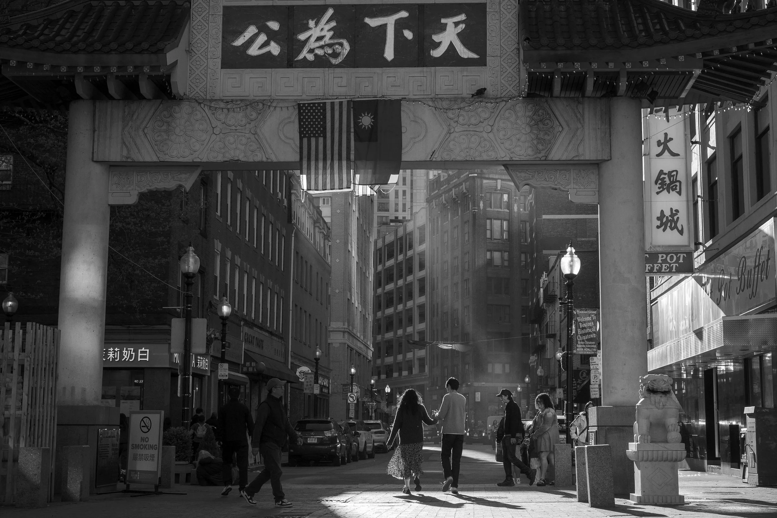 A street scene in Chinatown with a traditional Chinese archway featuring Chinese characters, hanging flags, a row of parked cars, street lamps, and people walking, with tall buildings in the background.