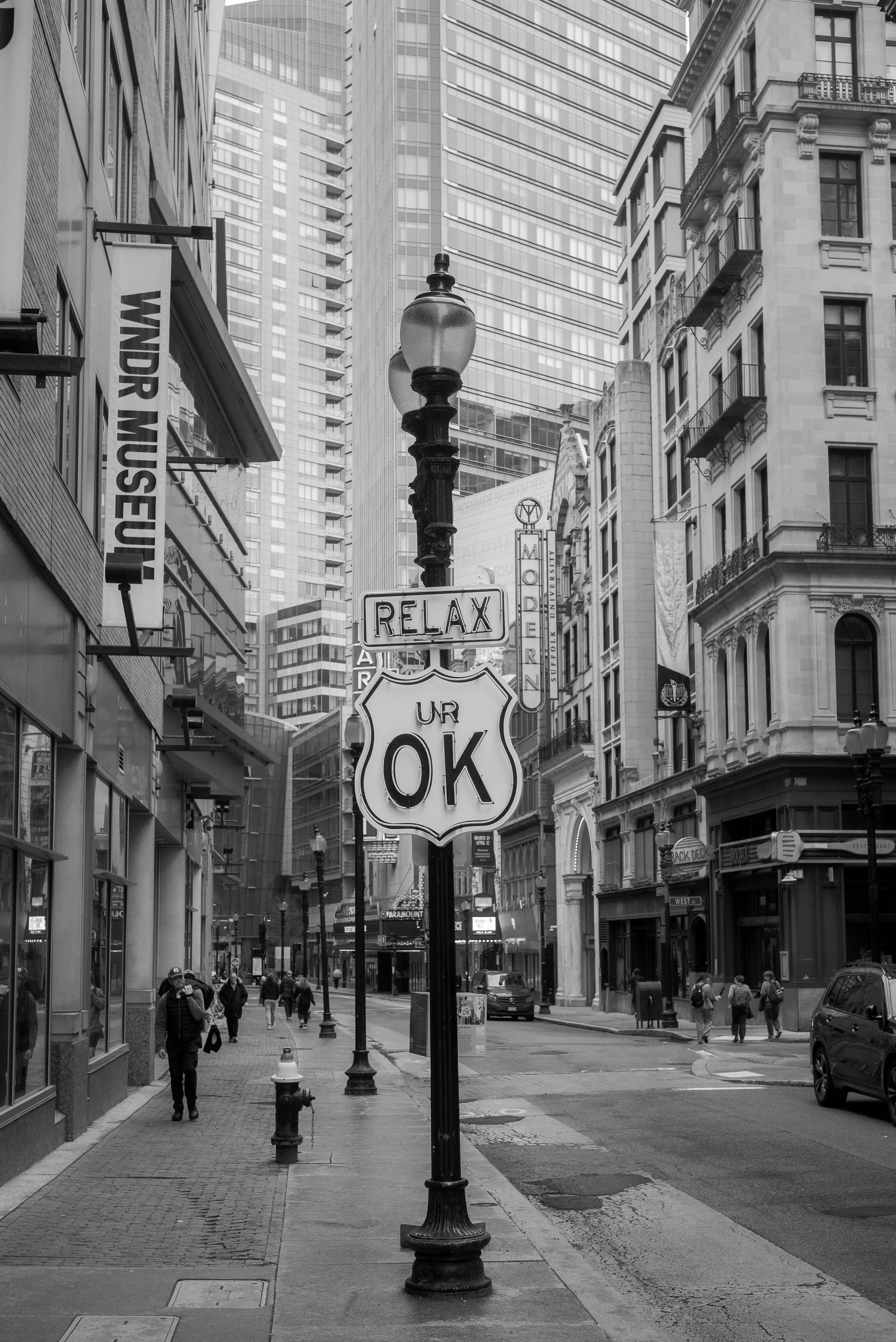 A black-and-white photo of a city street scene with tall buildings, a streetlamp, and pedestrians walking on the sidewalk. A sign reading 'Relax' is attached to the lamp post.
