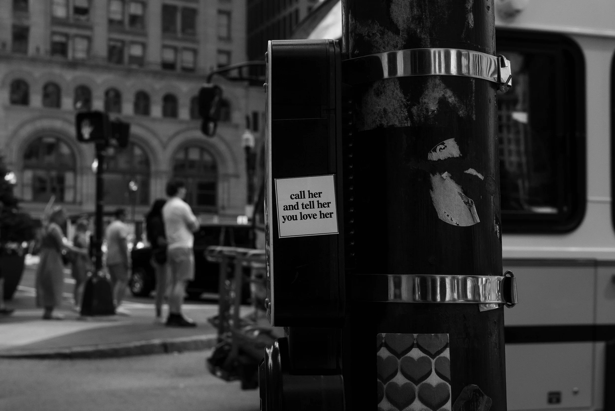 Close-up of a city street corner with a black pole featuring a sticker that says, 'call her and tell her you love her,' with blurred pedestrians and buildings in the background.