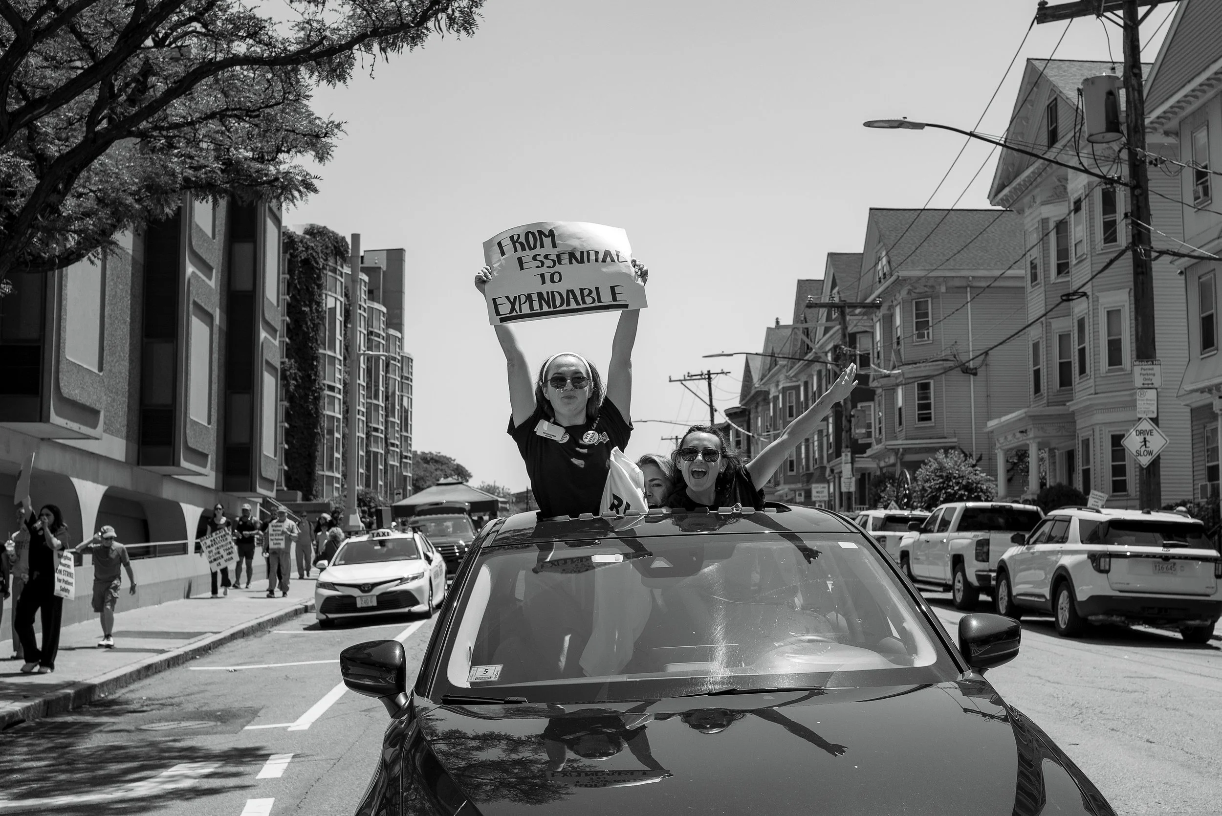 People participating in a parade on a city street, with one person holding a sign that reads "From Essential to Expensive" while standing through a car window. The street is lined with buildings, trees, and parked cars.