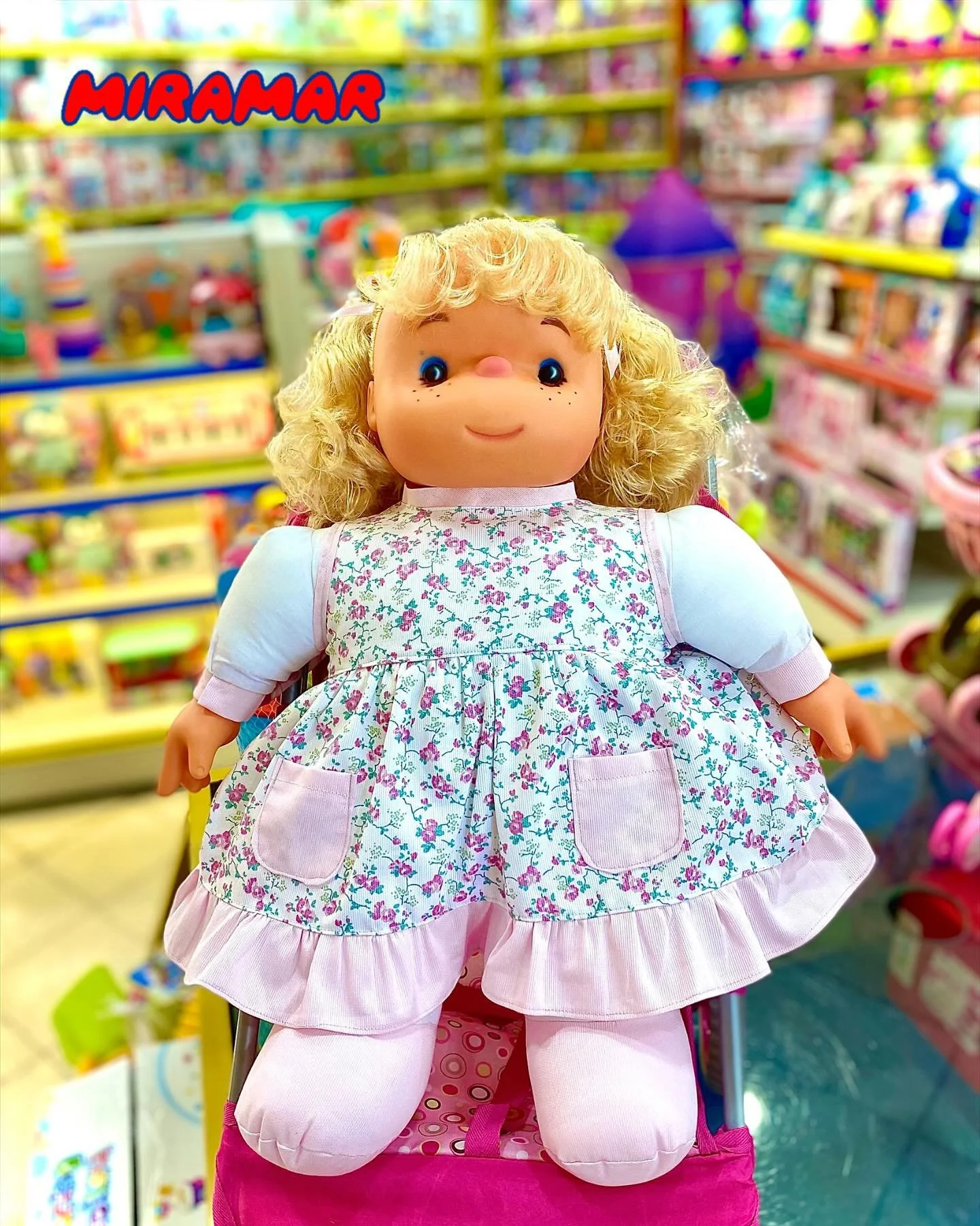 A large doll with curly blonde hair, blue eyes, and freckled cheeks, wearing a floral dress with pink pockets, sitting on a pink toy stroller in a colorful toy store aisle.