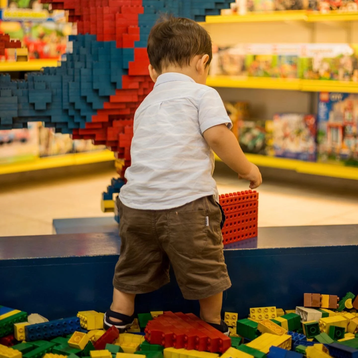 Young boy wearing a white shirt and brown shorts playing with large LEGO bricks in a toy store.